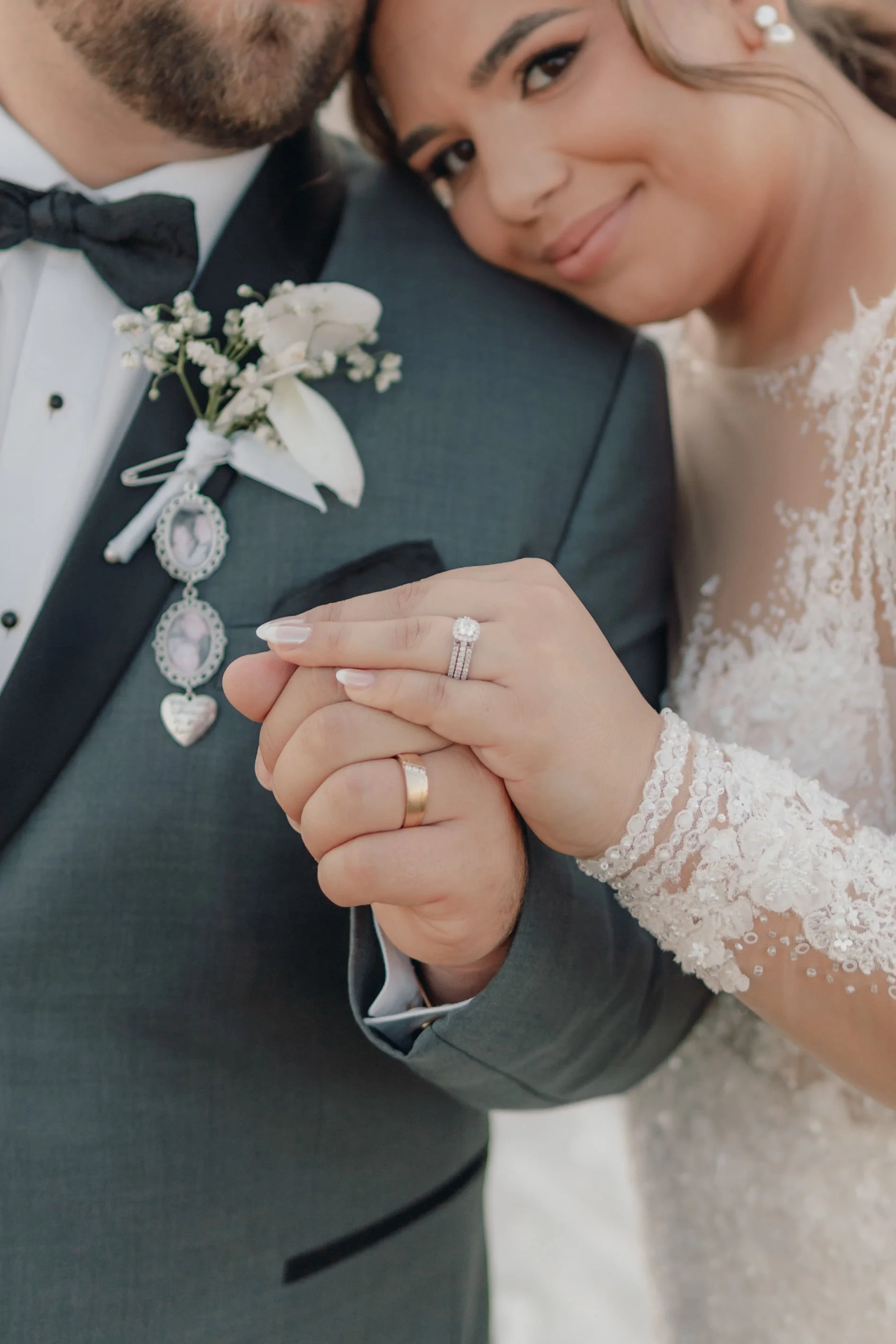 Close-up of a bride and groom holding hands, showing wedding rings, with the bride smiling and leaning her head on the groom's shoulder. The groom is wearing a suit with a boutonniere, and the bride is in a lace wedding dress.