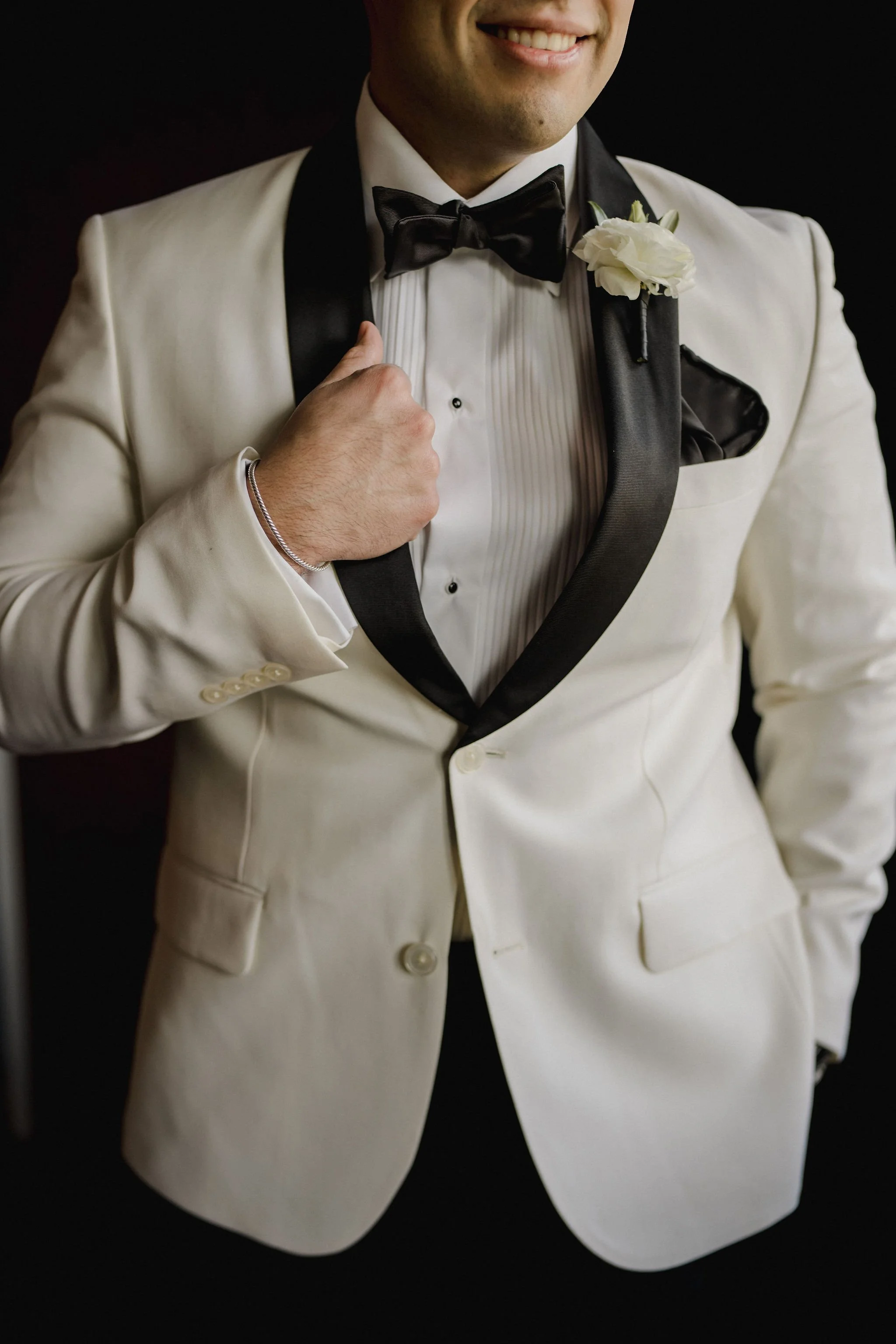 A man wearing a white tuxedo with black lapels, black bow tie, and a white shirt with pleats, giving a thumbs-up gesture with his right hand. The tuxedo jacket has a white flower boutonniere on the left lapel.