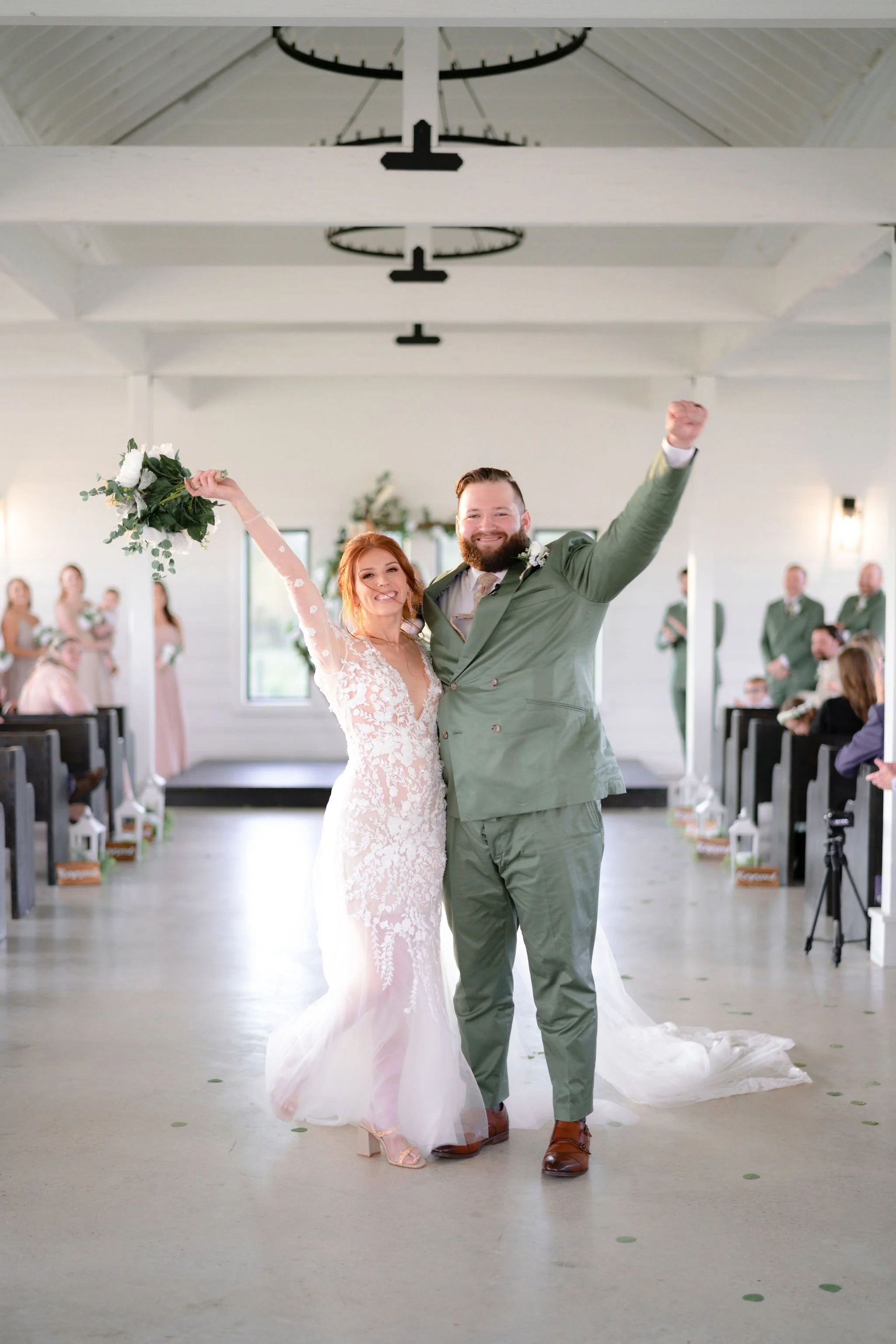 A bride and groom celebrating their wedding victory, with the bride holding a bouquet and both smiling, inside a bright, white-walled ceremony hall with guests in the background.