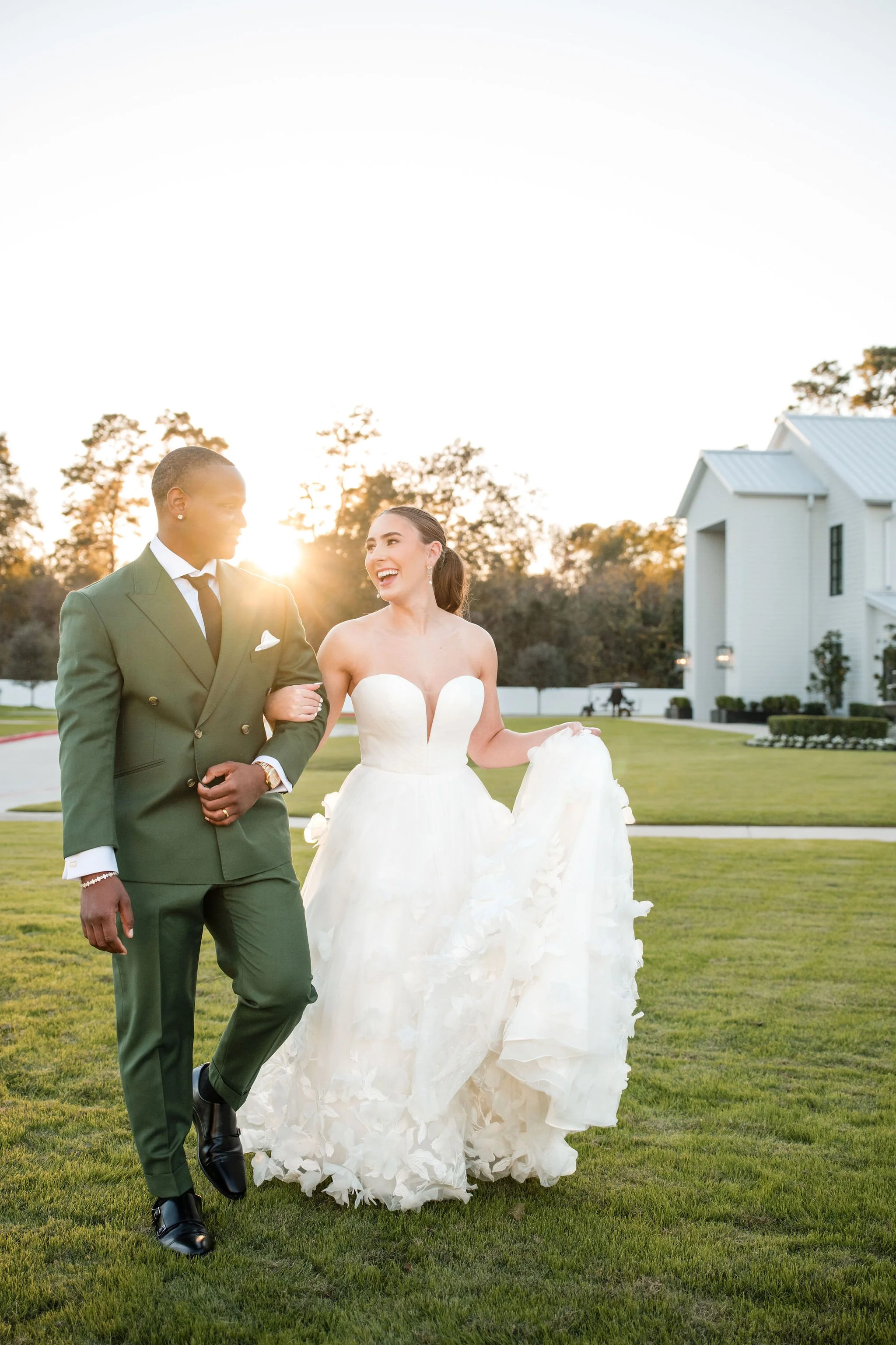 A bride in a white wedding dress and a groom in a green suit walk together on a grassy lawn during sunset, smiling and enjoying their moment.