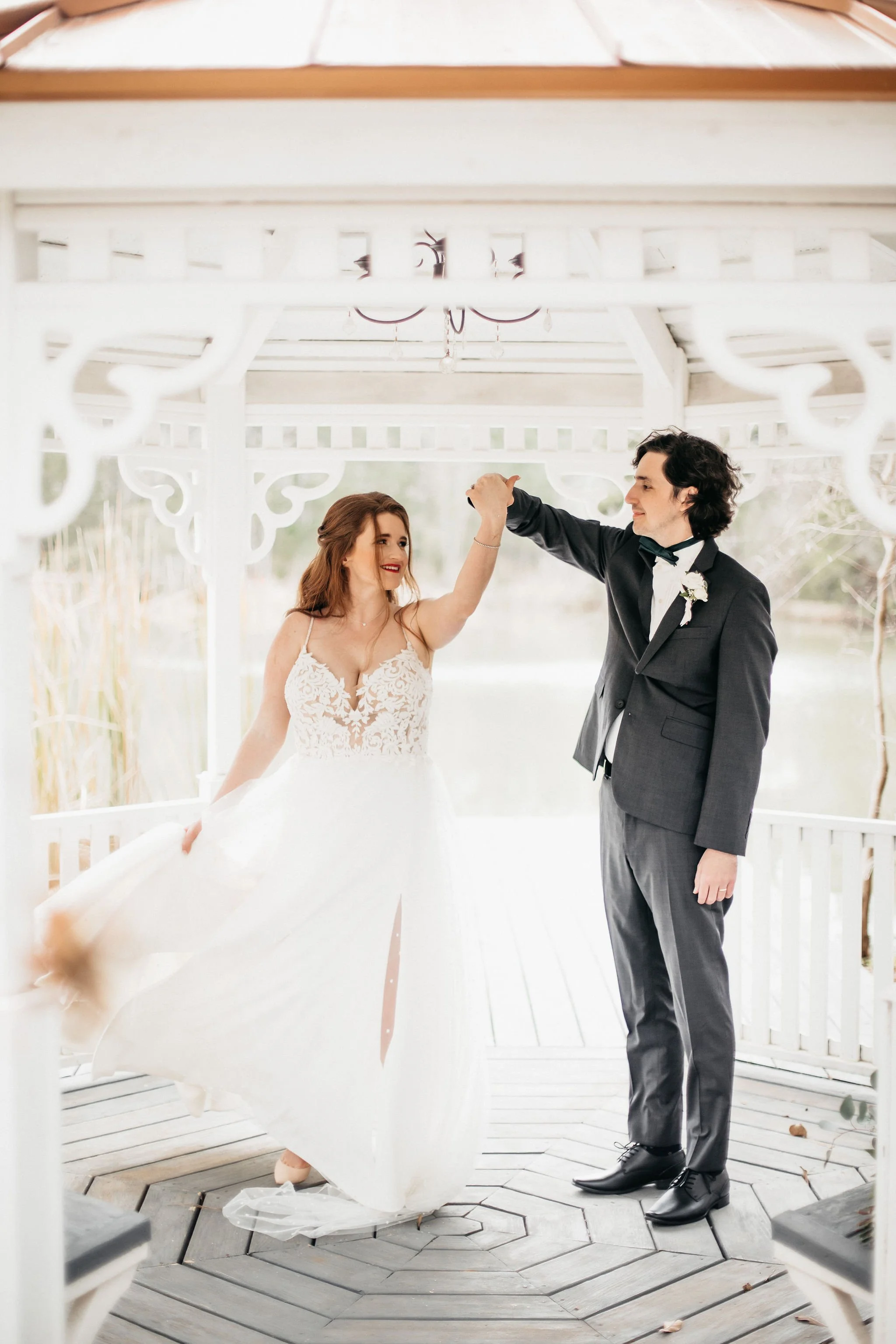 A bride and groom dancing together on a white wooden gazebo near a body of water, with the groom lifting the bride's hand.