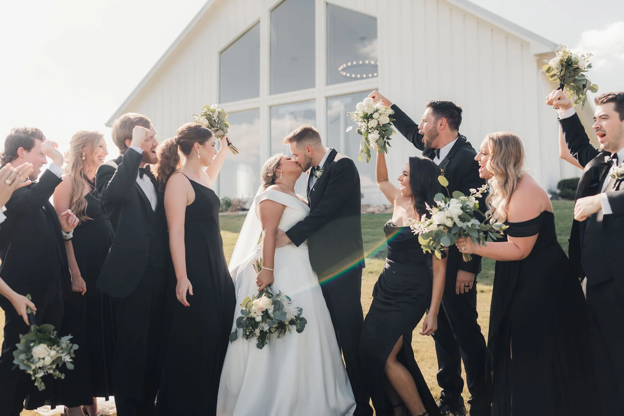Bride and groom kissing surrounded by wedding party celebrating outside a modern white building in sunlight.