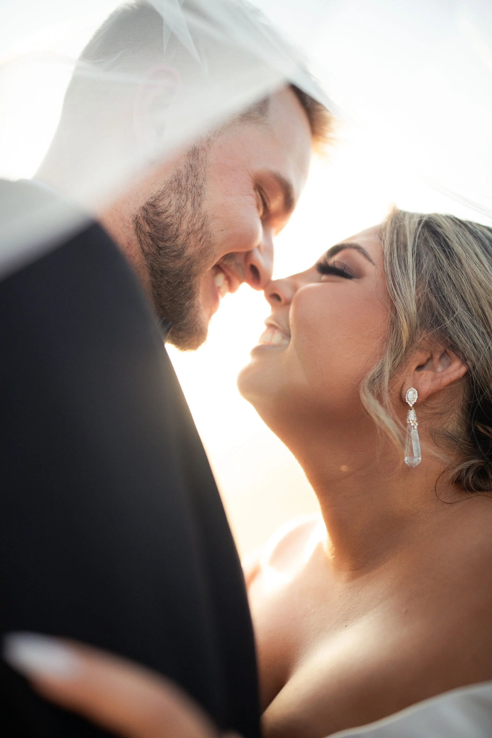 A close-up of a bride and groom smiling with noses touching, outdoors with bright sunlight.