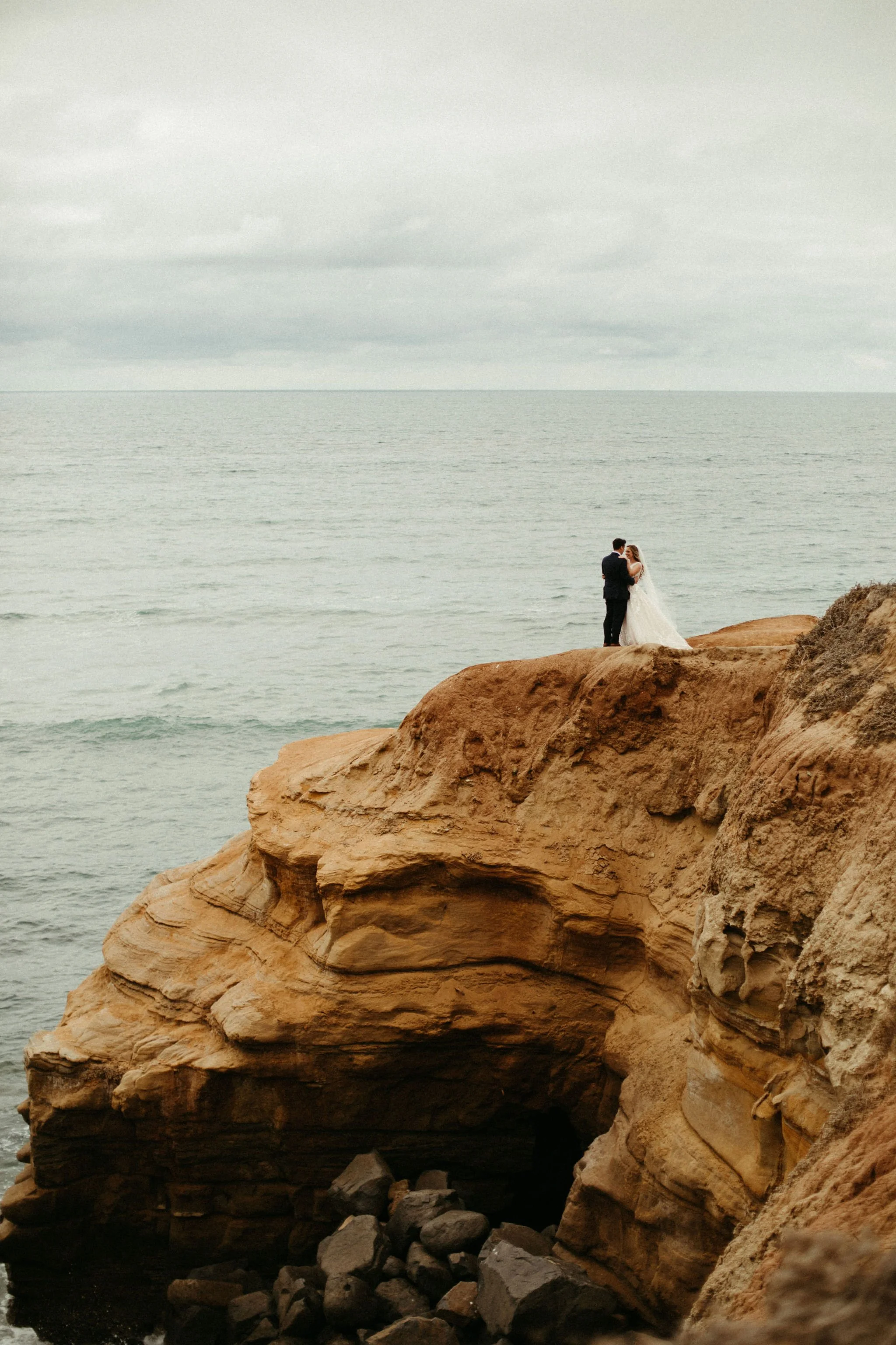 A bride and groom standing on a rocky cliff by the ocean.