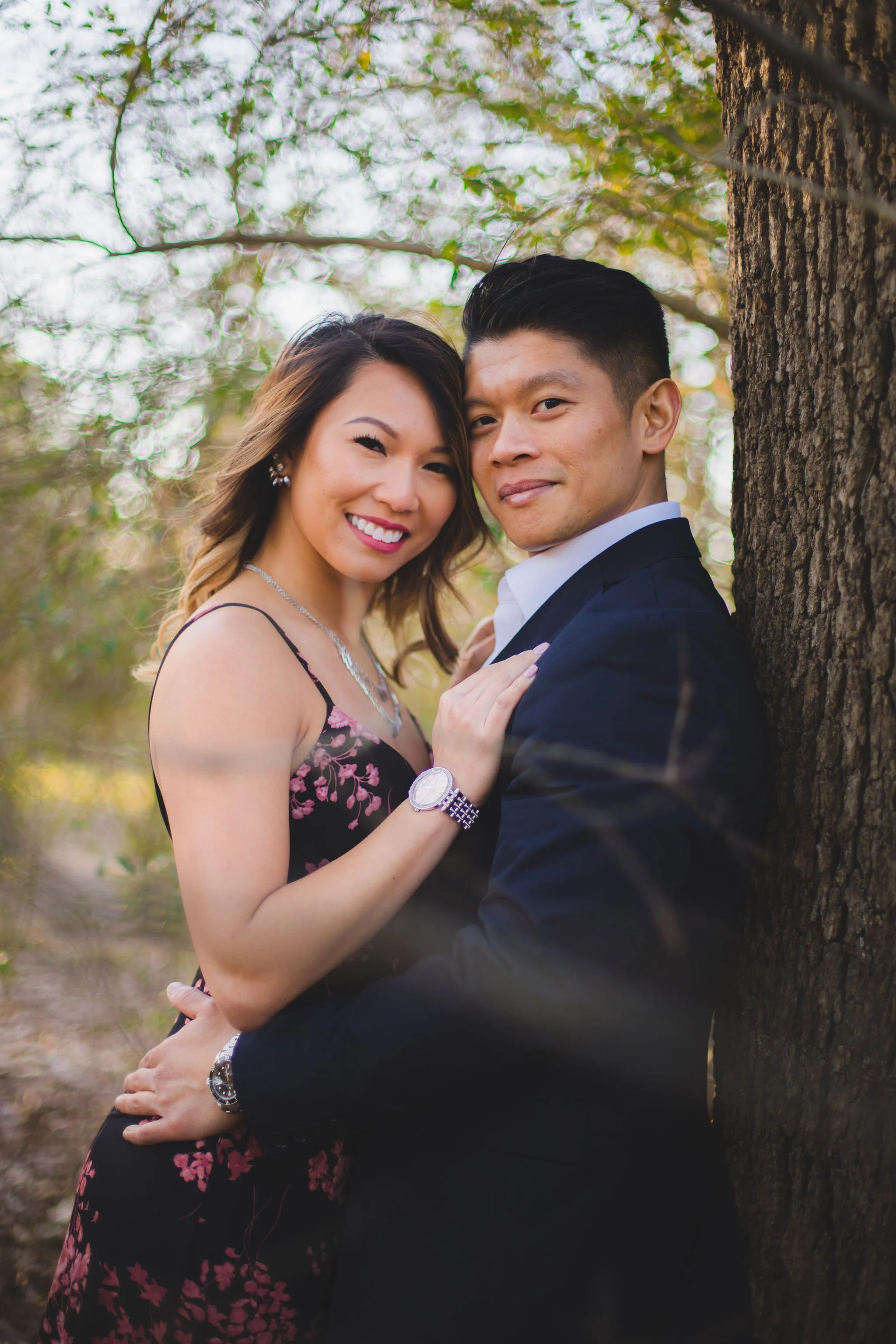 A smiling couple, a woman in a floral dress and a man in a black suit, standing close together outdoors next to a tree with blurred foliage in the background.