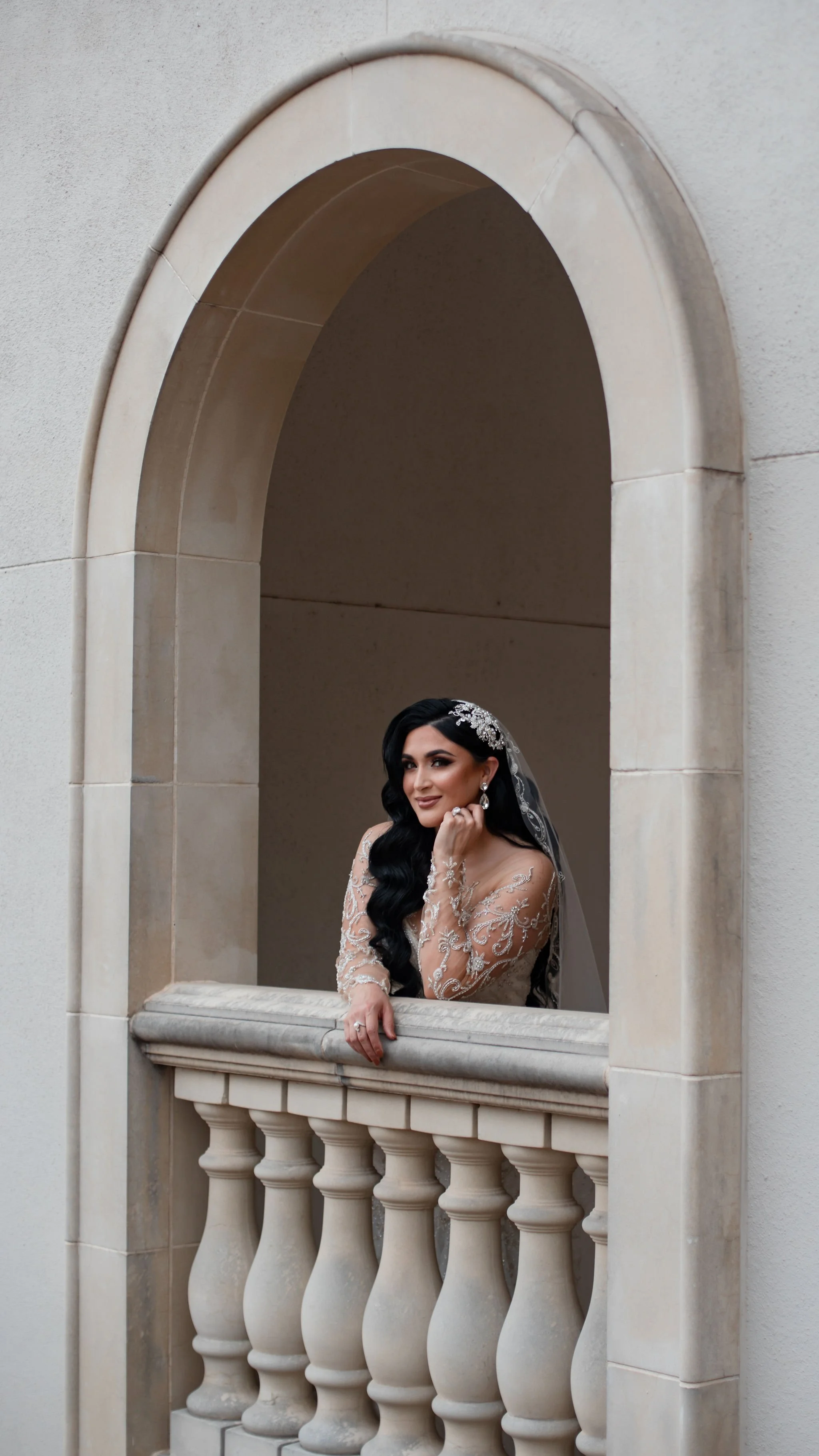 A bride with long dark hair and a lace wedding dress, standing on a stone balcony within an arched opening, smiling and holding her cheek with one hand.