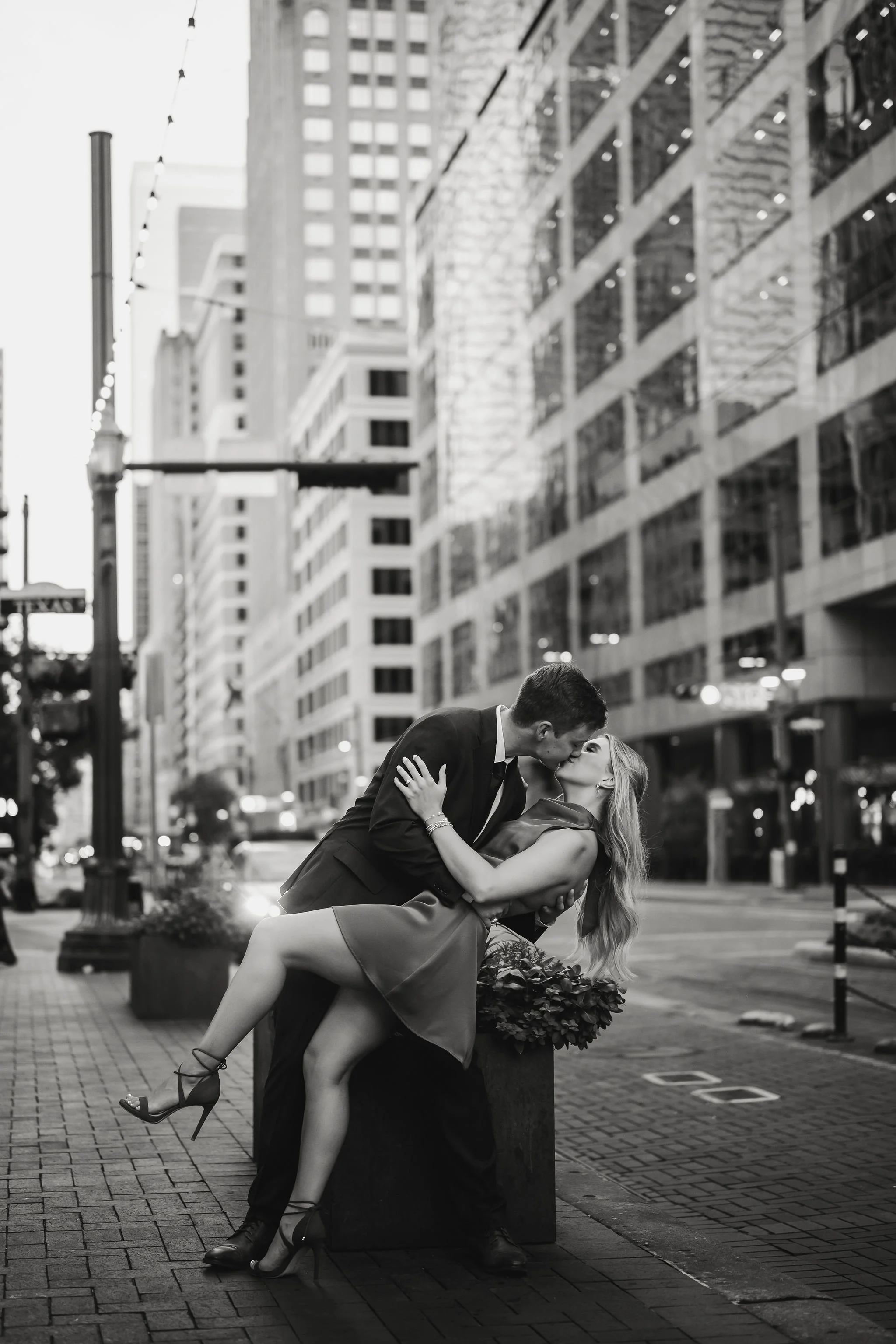 A black and white photograph of a passionate kiss between a man in a suit and a woman in a dress on an urban street with tall buildings.