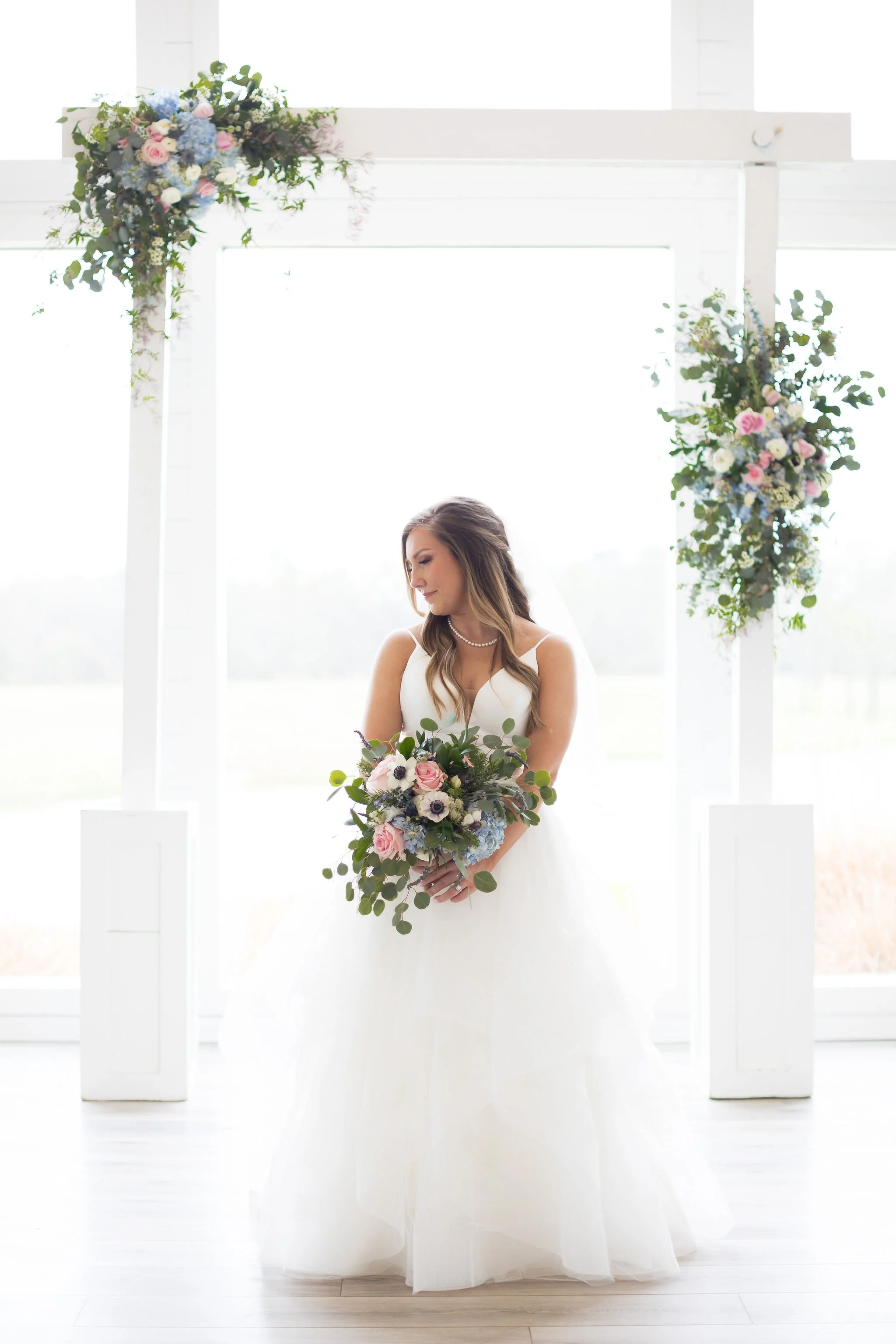 A bride in a white wedding gown holding a bouquet of flowers, standing in front of a decorated wedding arch with floral arrangements.