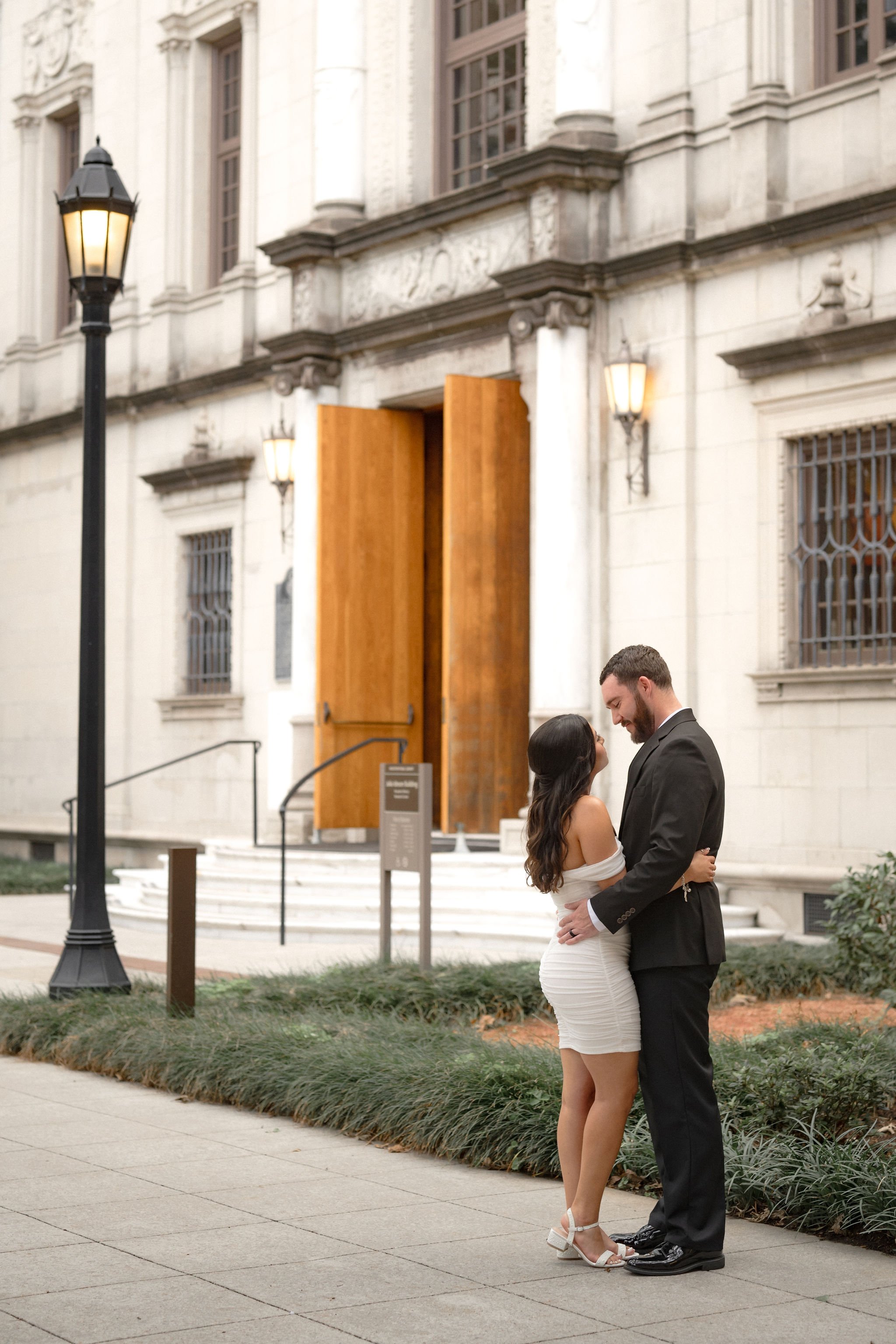 A couple dressed in formal attire sharing a romantic moment outside a historic building during the evening. The woman is wearing a white off-shoulder dress and white heels, and the man is in a black suit and tie. The background features a large woode