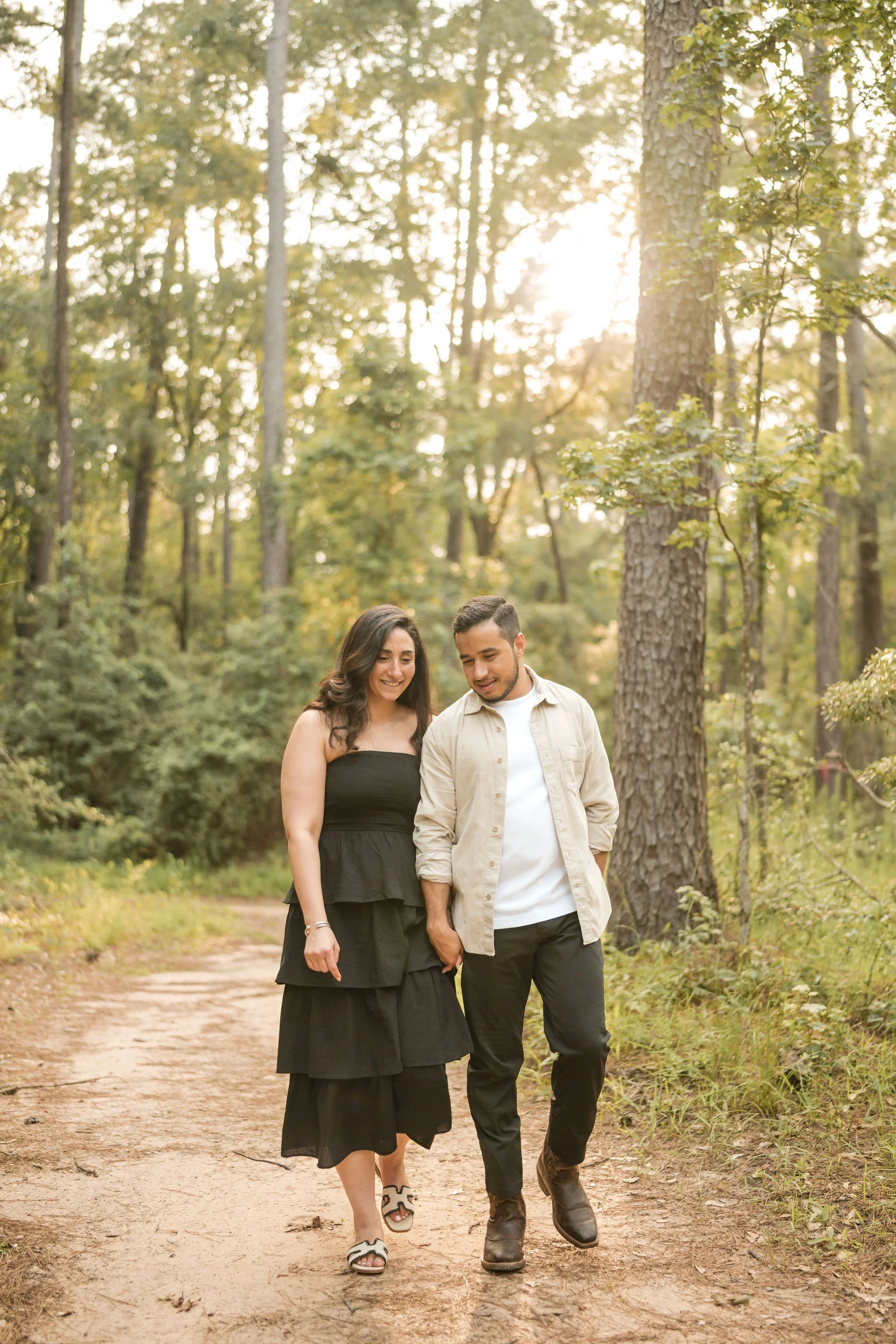 A couple walking hand in hand on a dirt trail through a forest, smiling and enjoying the outdoors during what appears to be late afternoon or early evening.
