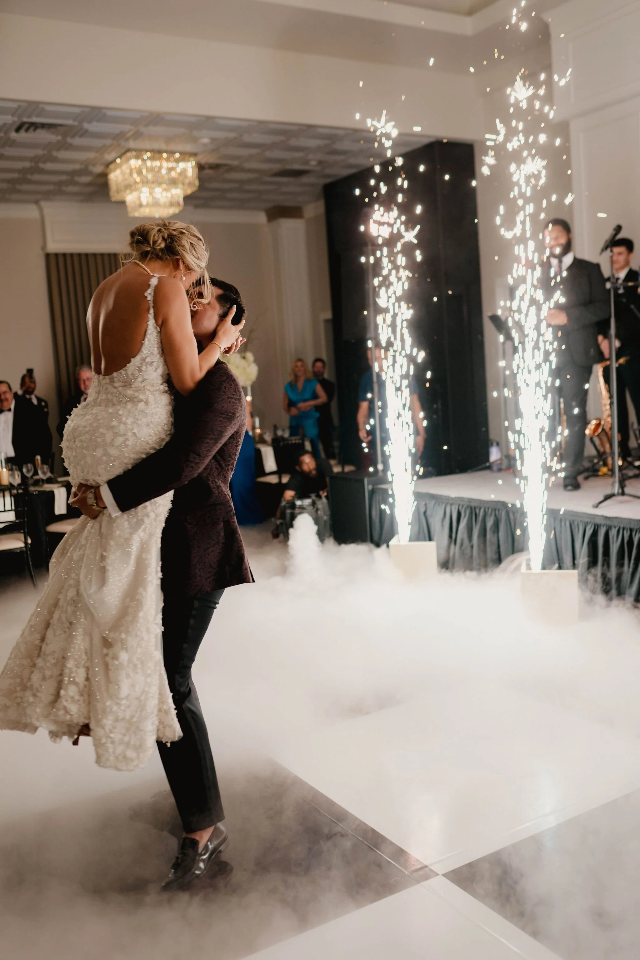 A bride being lifted by a groom during a wedding celebration with sparklers and smoke effects.