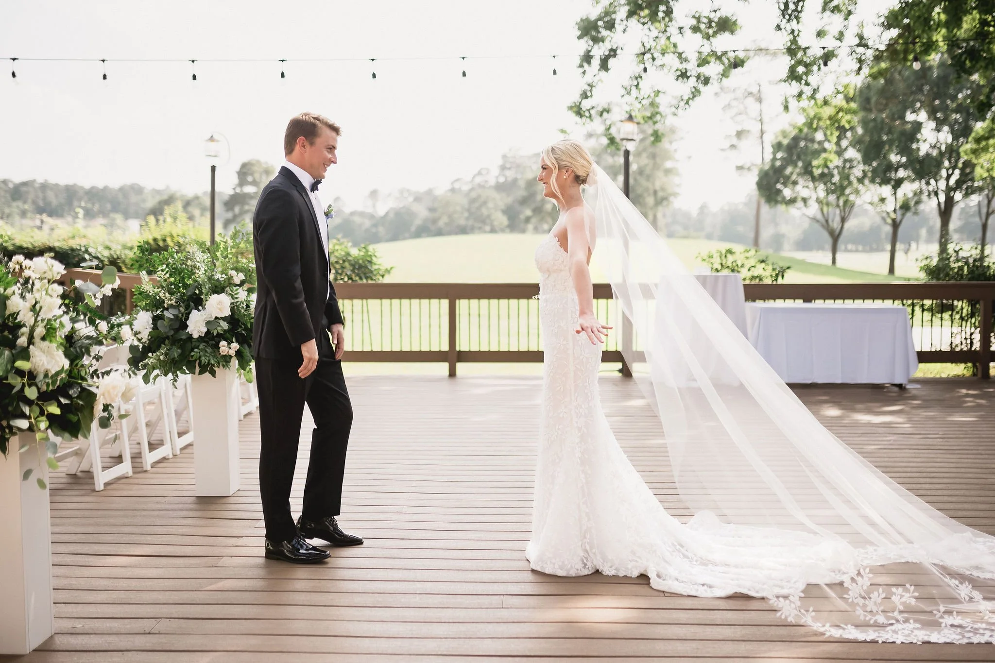 A bride and groom on a wooden deck during their outdoor wedding ceremony, with the bride in a white lace gown and veil, and the groom in a black tuxedo, smiling at each other amid floral arrangements and greenery.