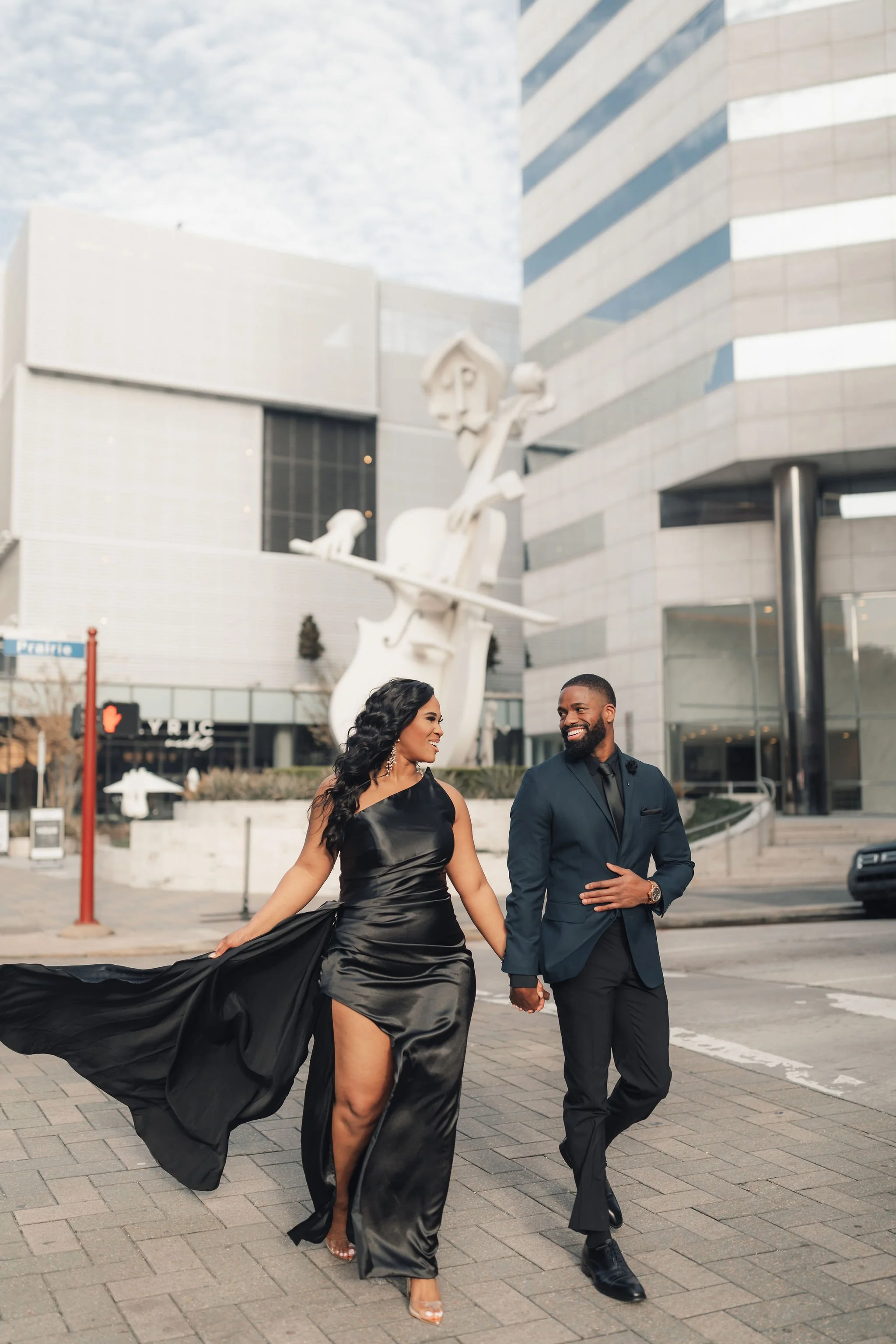 A smiling couple, woman in a black satin gown and man in a navy suit, holding hands and walking outdoors in front of a modern building with a large abstract sculpture of a person playing a string instrument.