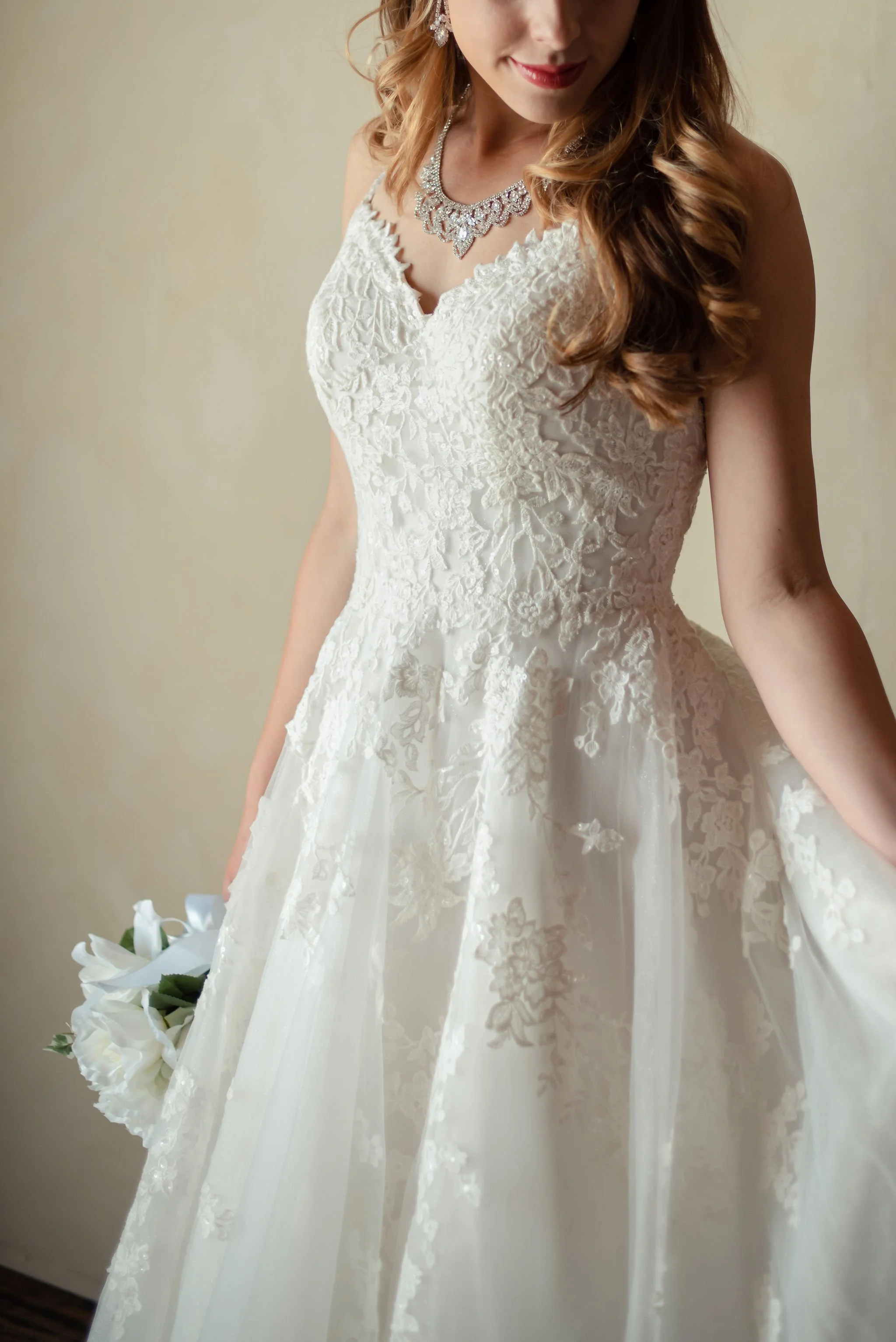 A woman in a white wedding dress, holding a bouquet of white flowers, with jewelry including a necklace and earrings, and styled with loose curled hair.