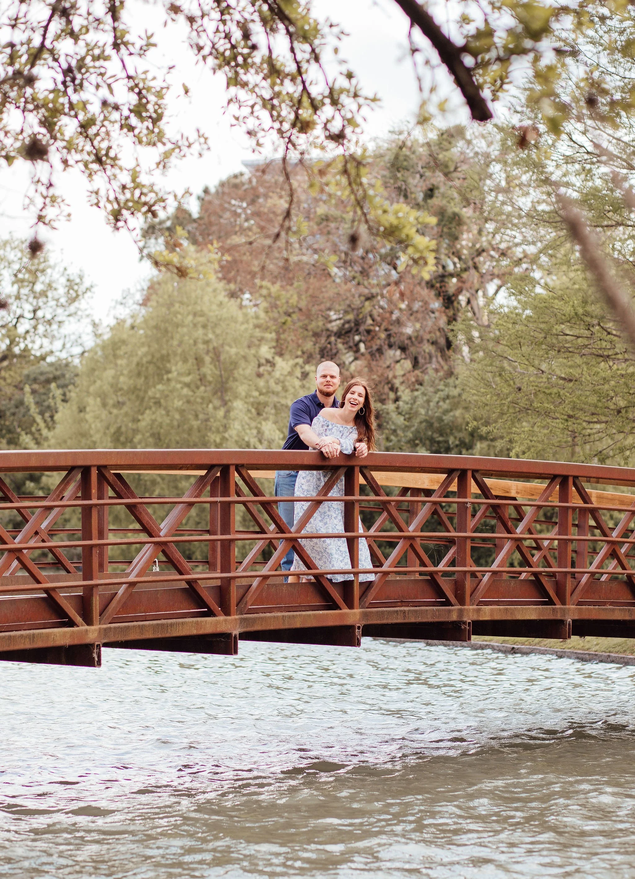 A couple stands on a wooden bridge over a body of water, smiling and embracing in a park with trees and greenery in the background.