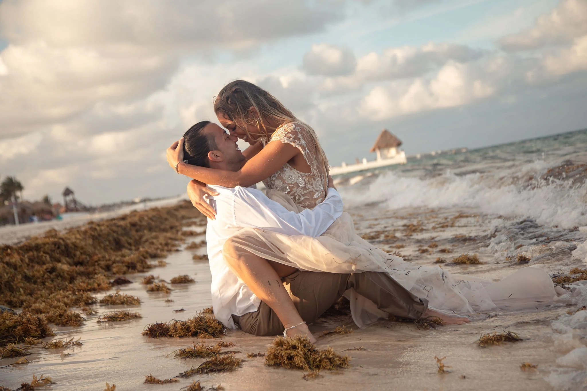 A couple in wedding attire embracing on a beach with seaweed, waves, and a thatched-roof hut in the background.
