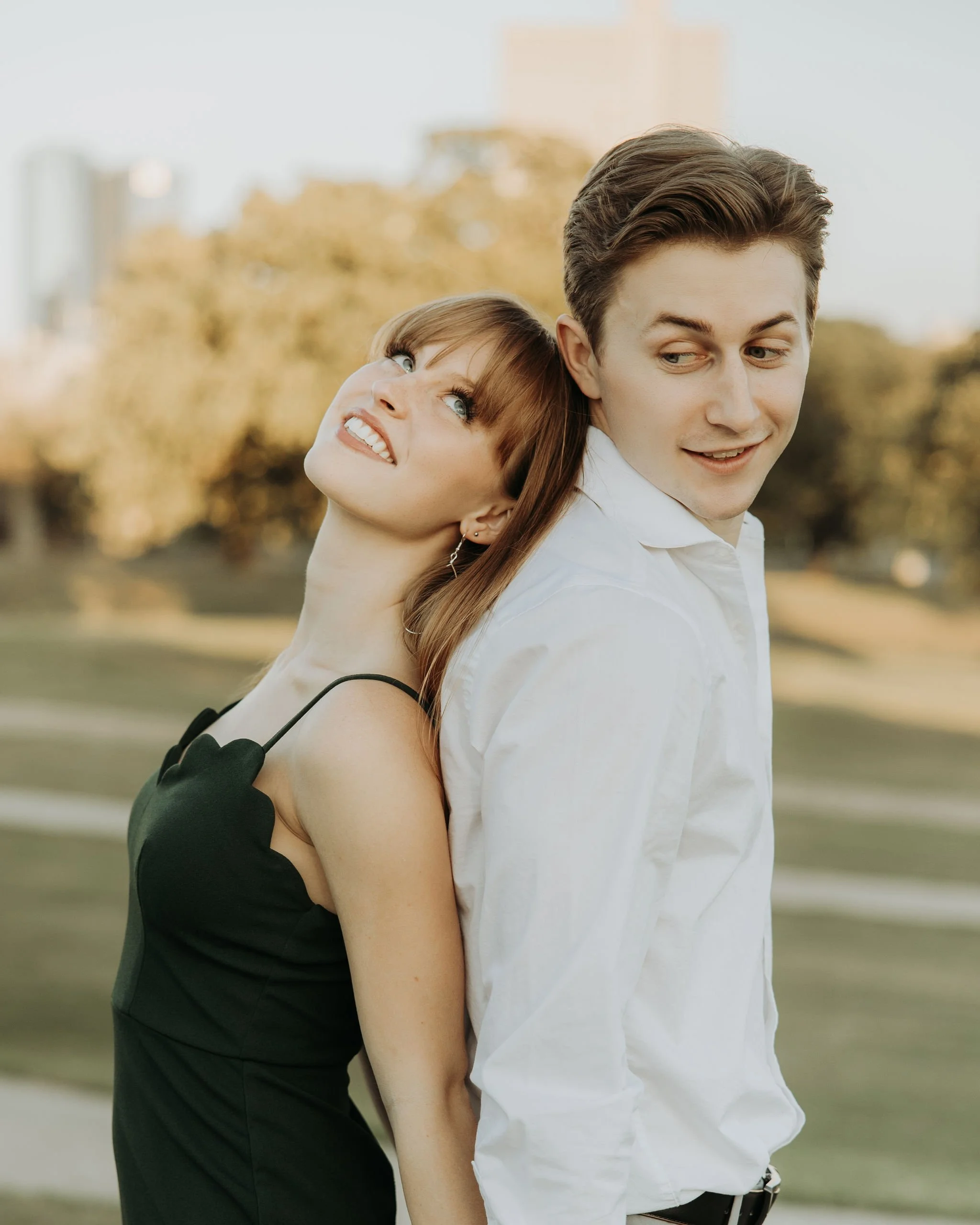 A woman and a man standing back to back outdoors, woman making a playful grimace and the man looking to his right with a slight smile, with trees and buildings in the background.