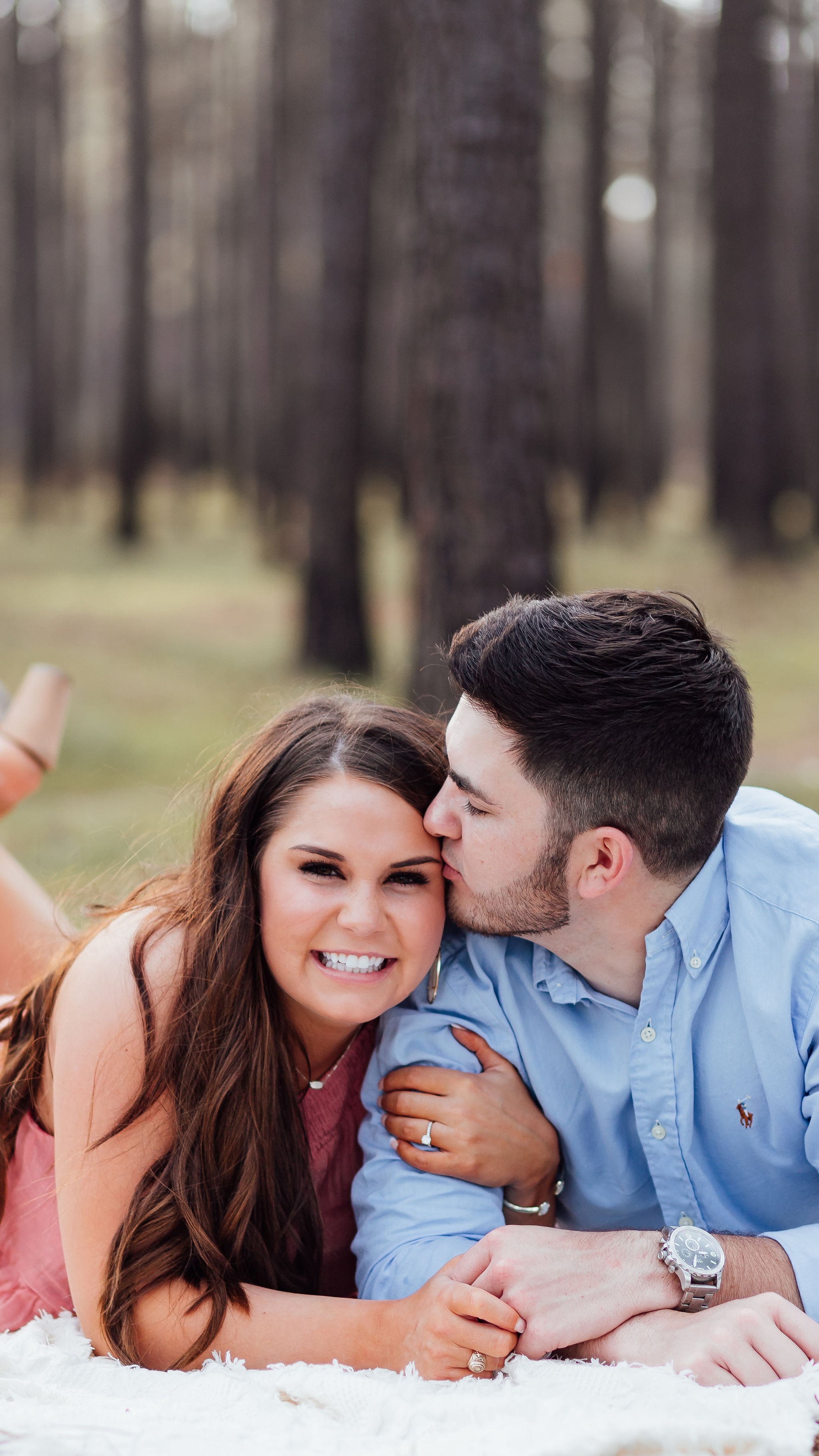 A couple lying on a blanket outdoors in a wooded area; the woman smiling and the man kissing her on the cheek.