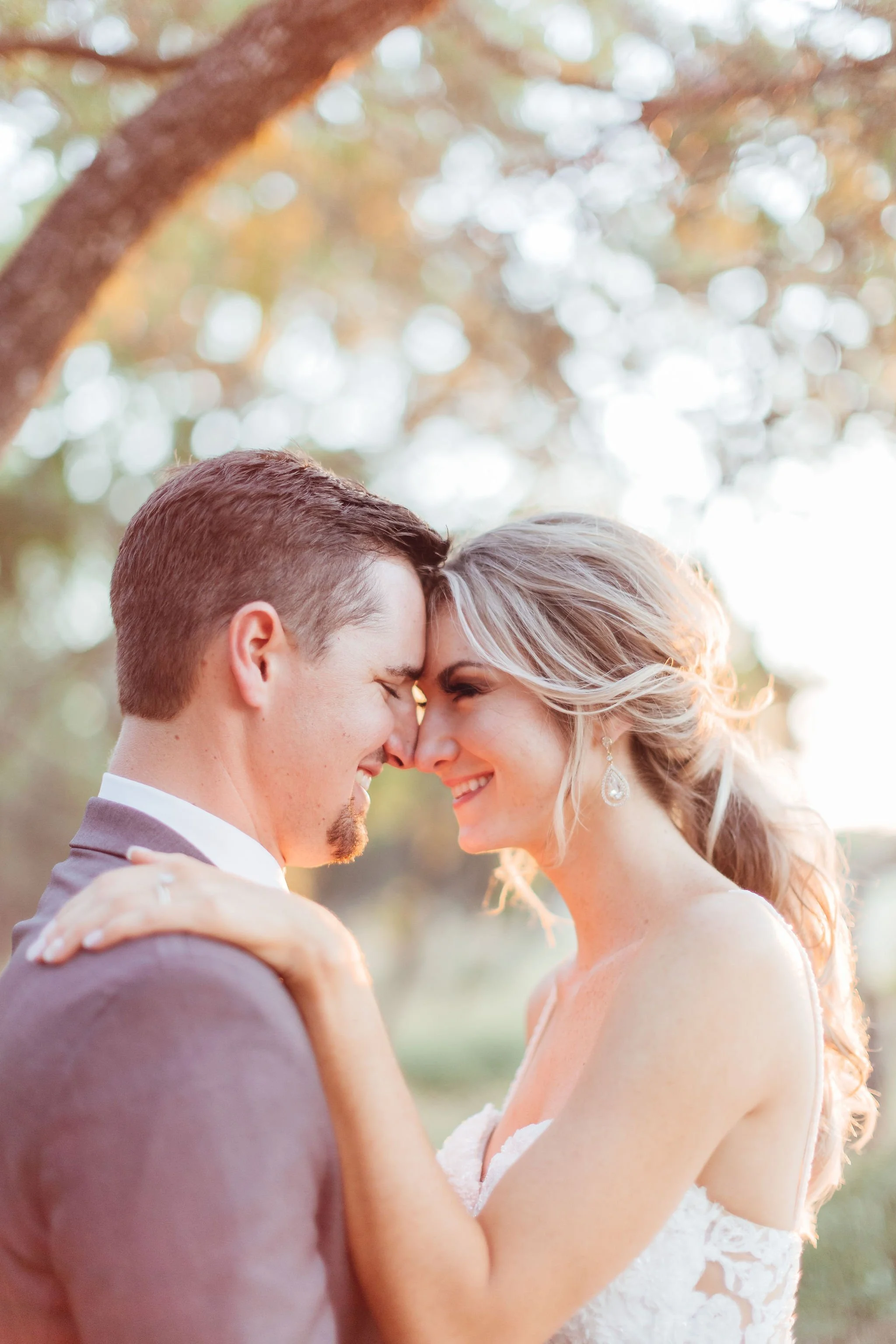 A groom and bride with foreheads touching, smiling, outdoors during sunset with blurred trees in the background.