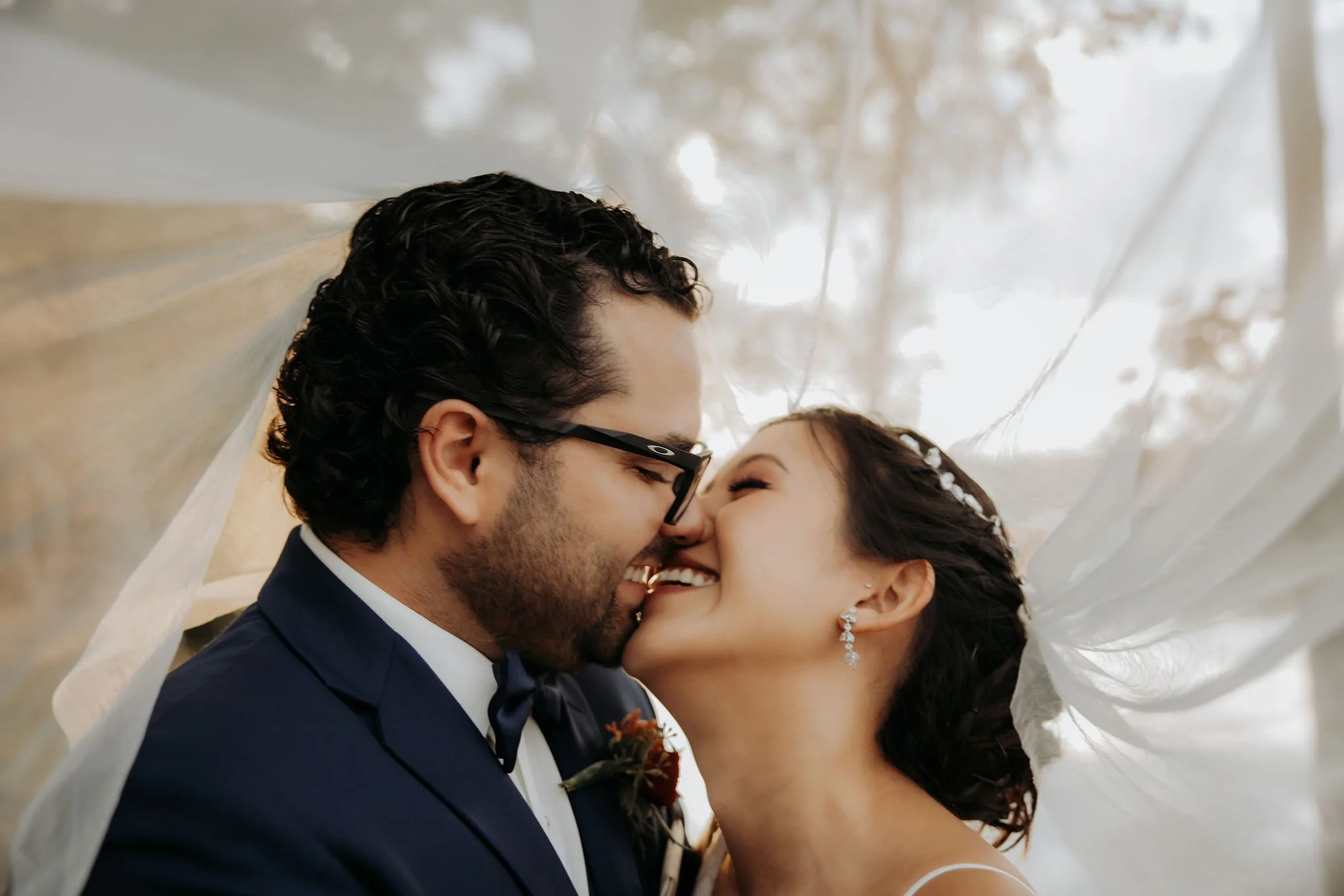 Close-up of a happy bride and groom sharing a kiss under a wedding veil, outdoors with trees in the background.