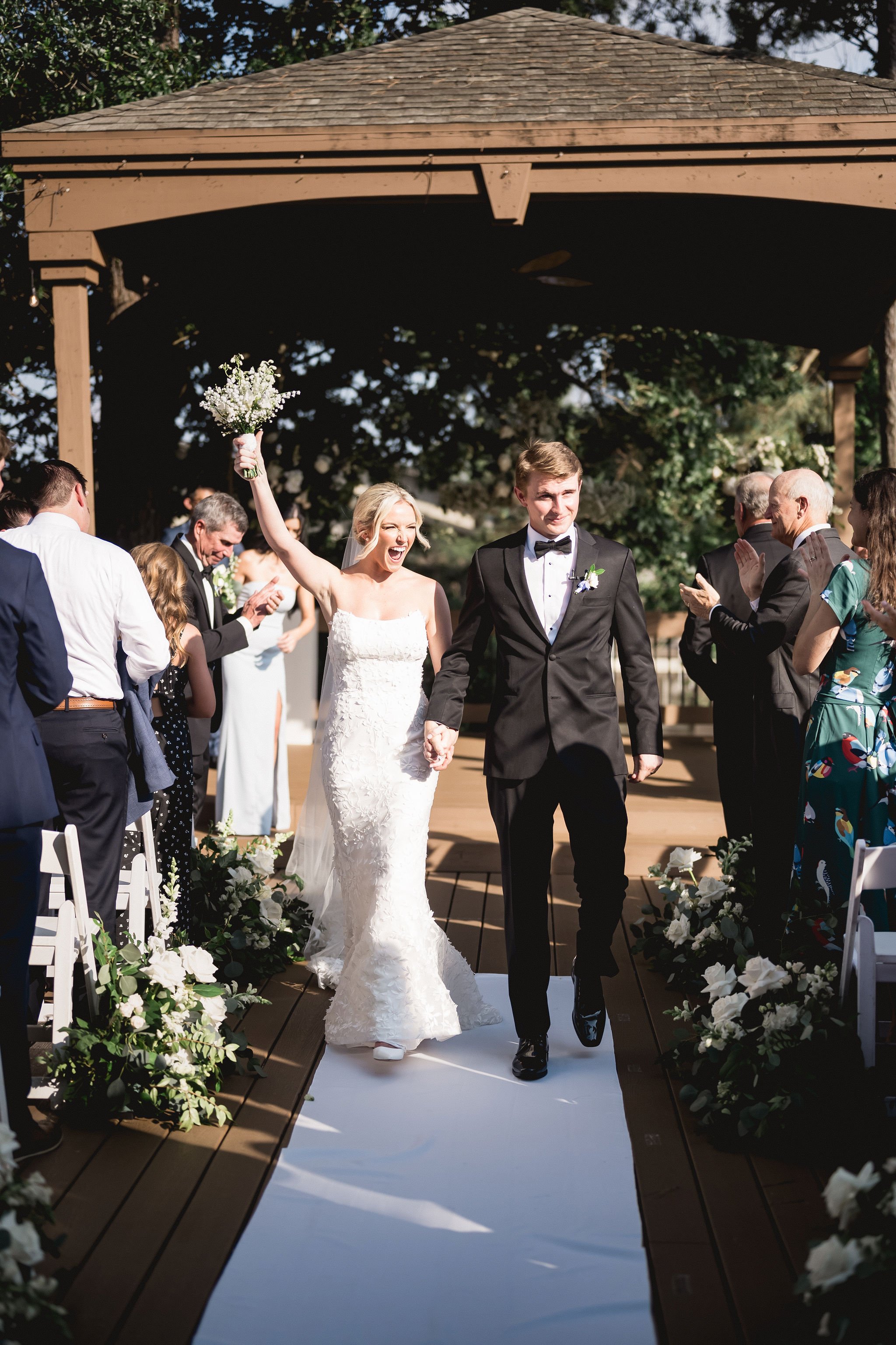 A newlywed couple walking down the aisle, holding hands, with the bride raising a bouquet in celebration while smiling, as guests clap and cheer on a sunny outdoor wedding ceremony on a decorated wooden pathway.