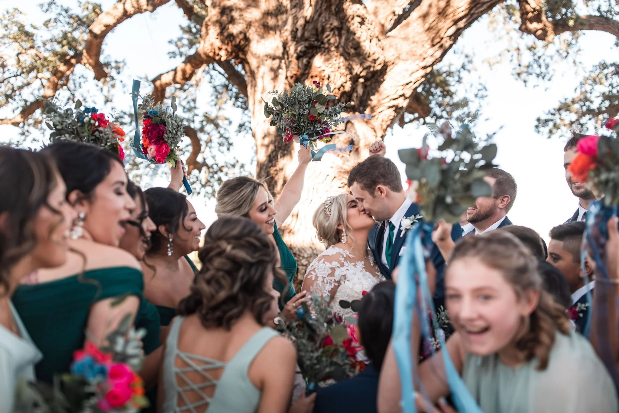 A wedding celebration outdoors with the bride and groom kissing under a large tree, surrounded by friends and family holding bouquets and smiling.