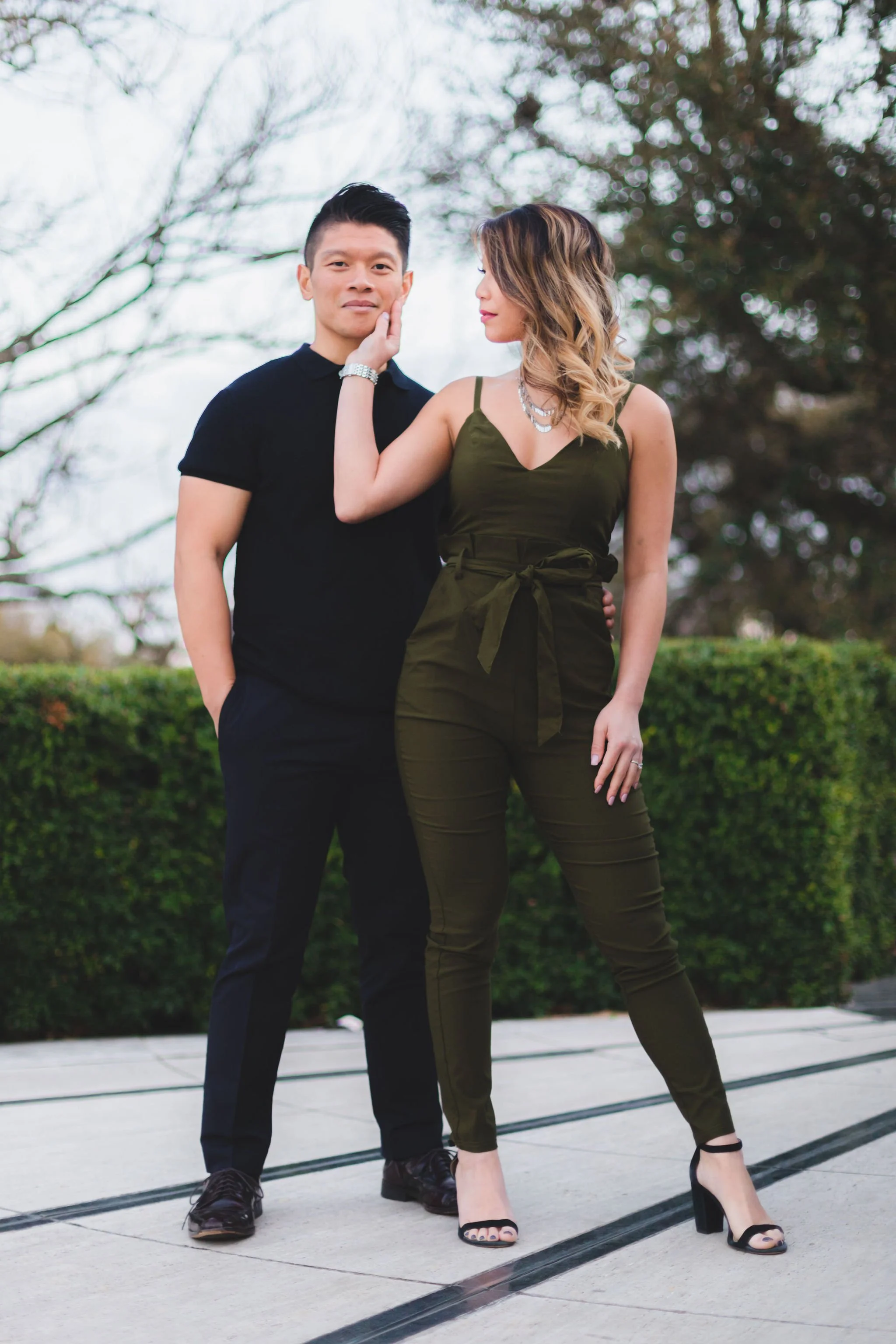 A man and woman standing outdoors on a paved sidewalk with greenery and trees in the background. The man has short dark hair and is wearing a black shirt and black pants. The woman has shoulder-length wavy hair, a sleeveless olive-green jumpsuit with