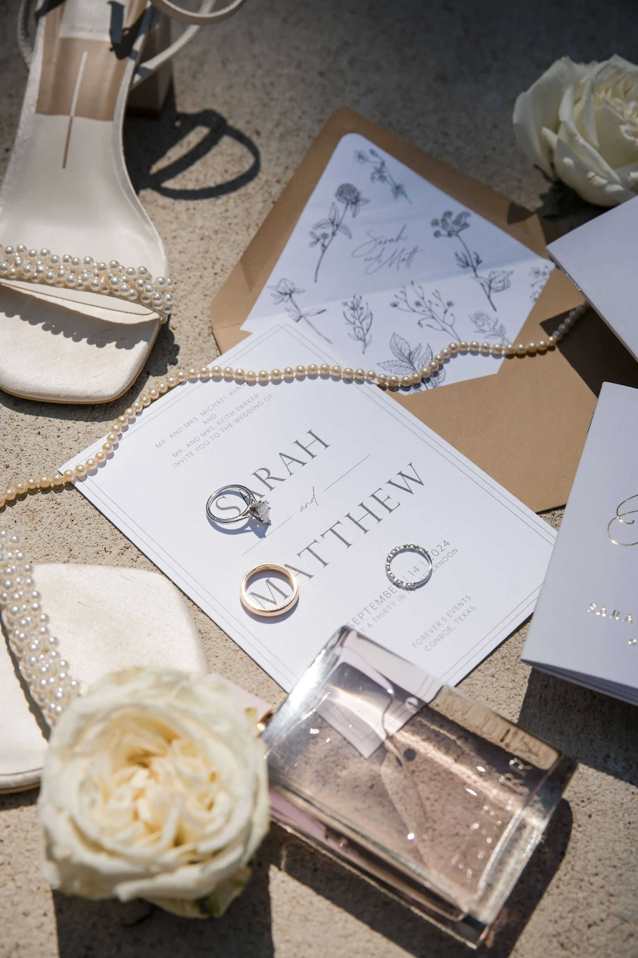 Wedding invitation with rings and accessories on sand, including pearl jewelry, white shoes with pearl embellishment, a white flower, and sketches of flowers.