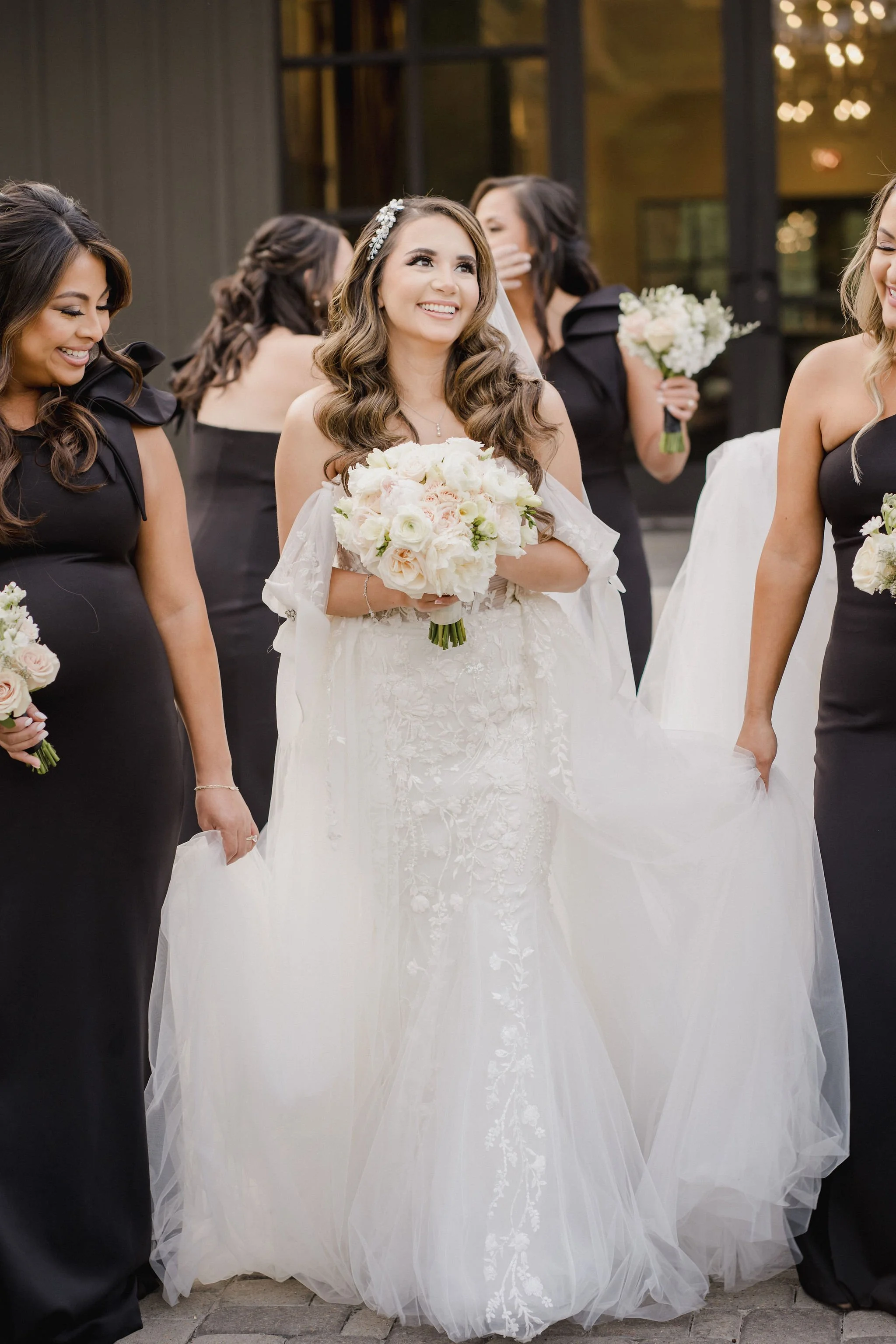 Bride in a white wedding gown holding a bouquet of white and pink flowers, surrounded by bridesmaids in black dresses, outdoors.
