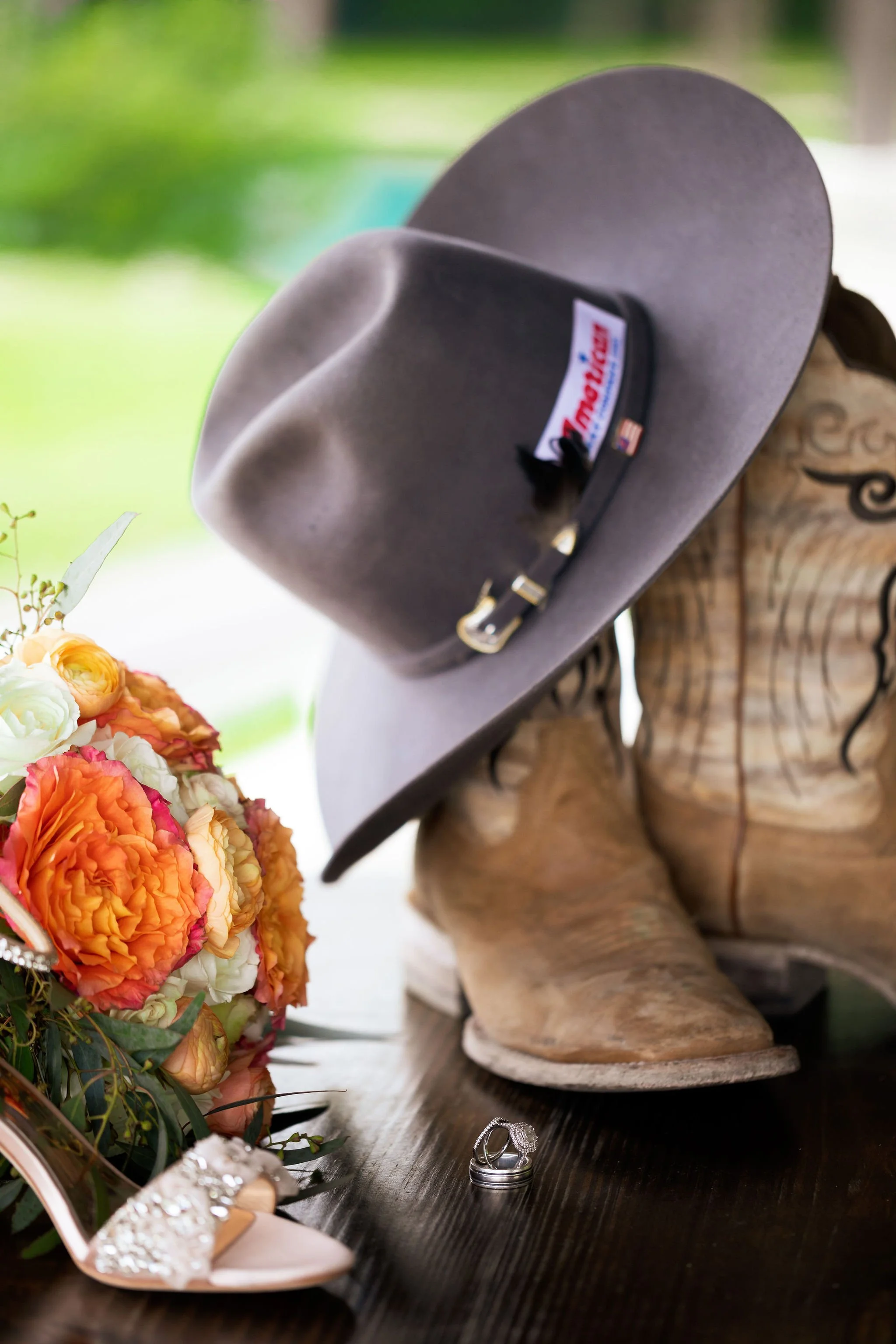 A cowboy hat, a bouquet of colorful flowers, a pair of wedding rings, and a high heel shoe with embellishments on a dark wooden table.