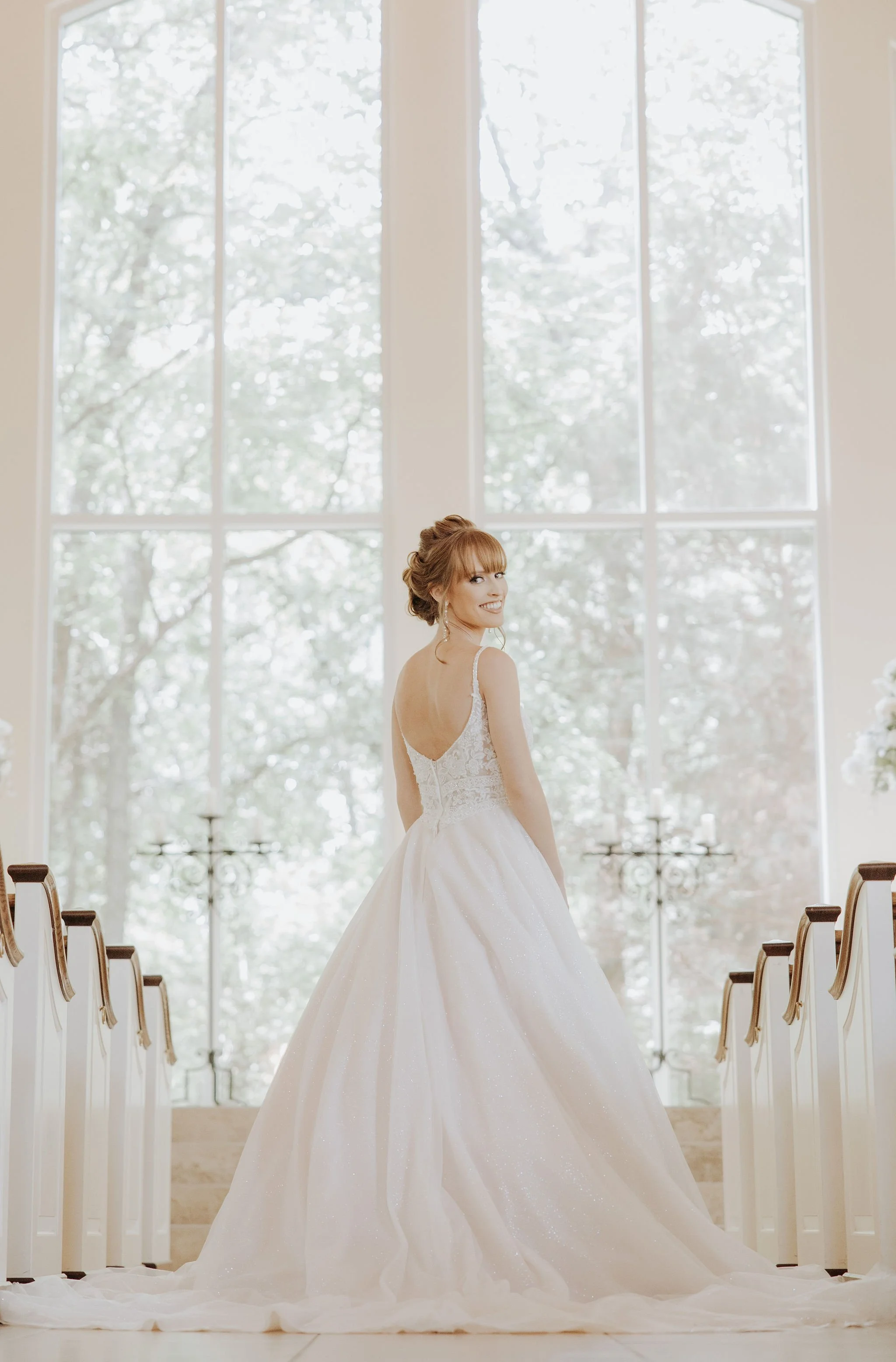 A bride in a white wedding gown standing inside a church, facing away but turning her head to smile at the camera, with large windows and natural light in the background.