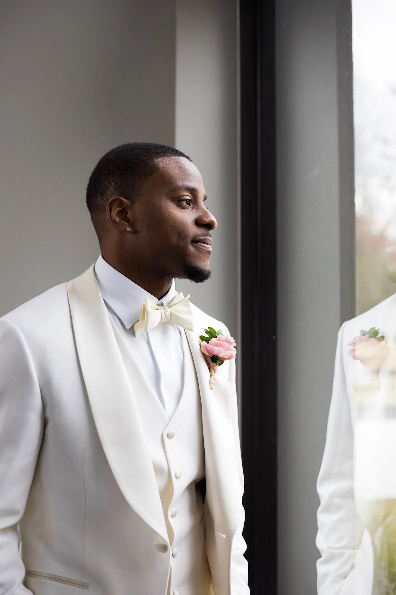 A groom in a white tuxedo with a bow tie and boutonniere, standing by a window, looking contemplative.