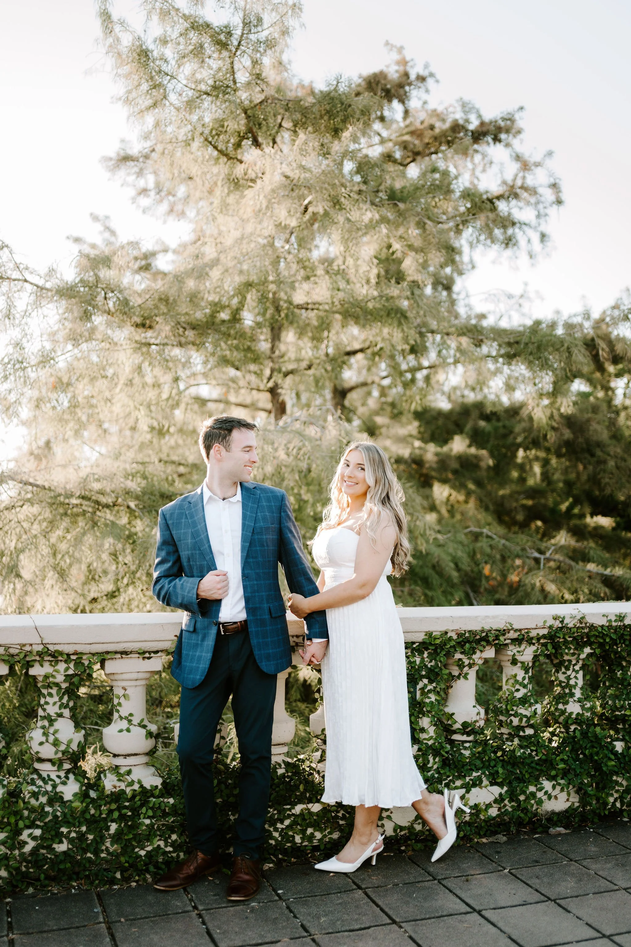 A smiling couple standing on a patio with a decorative white railing and greenery; woman in a white dress and heels, man in a blue plaid blazer and dark pants, holding hands and looking at each other in a natural outdoor setting with trees.