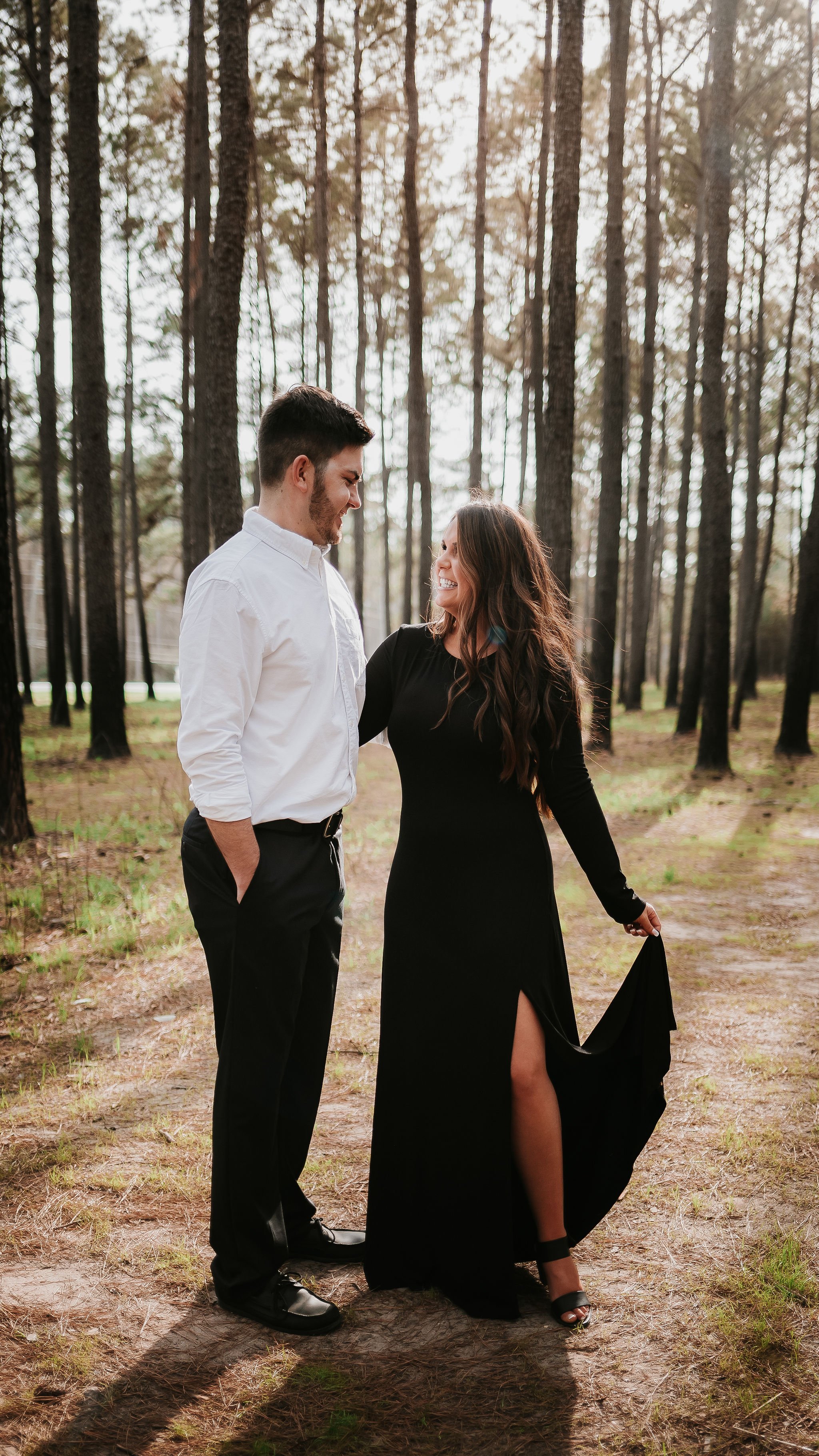 A man and woman standing close together in a forest, smiling at each other. The woman is wearing a long black dress with a slit, and the man is dressed in a white shirt and black pants. The sunlight filters through the trees.