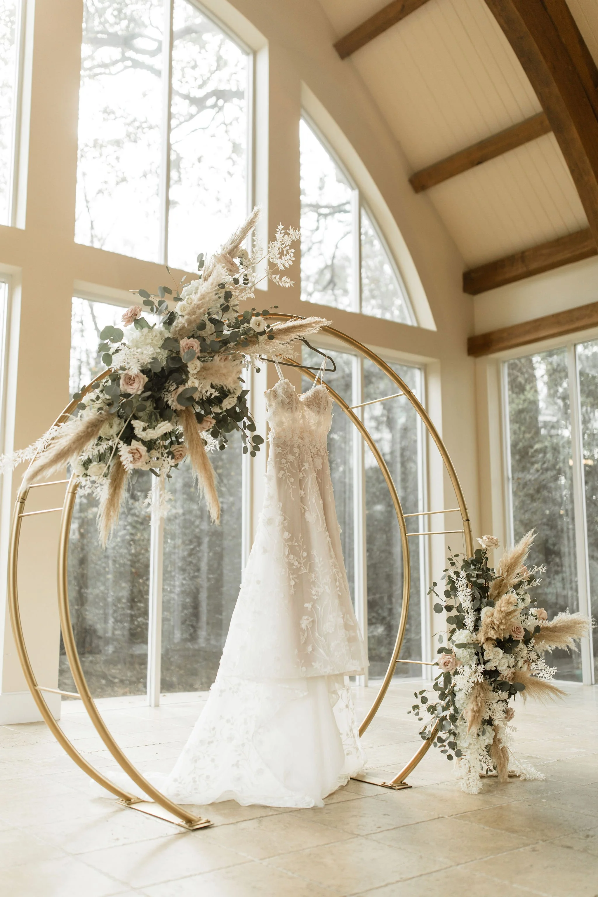 A white wedding dress hanging from a floral display on a gold circular arch in a bright, airy room with large windows and wooden beams.