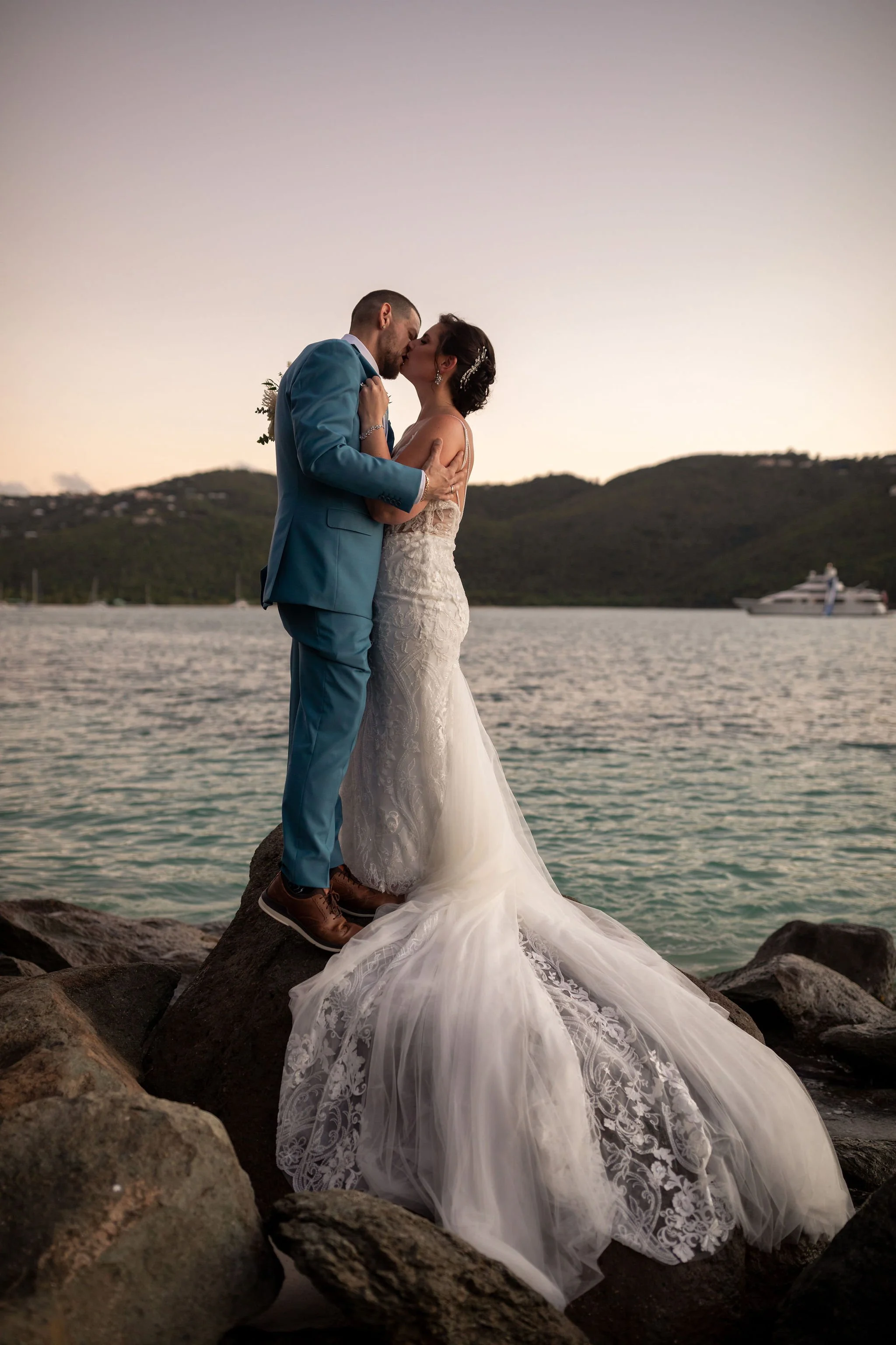 A bride and groom sharing a kiss on a rocky shoreline at sunset, with hills and a boat in the background.