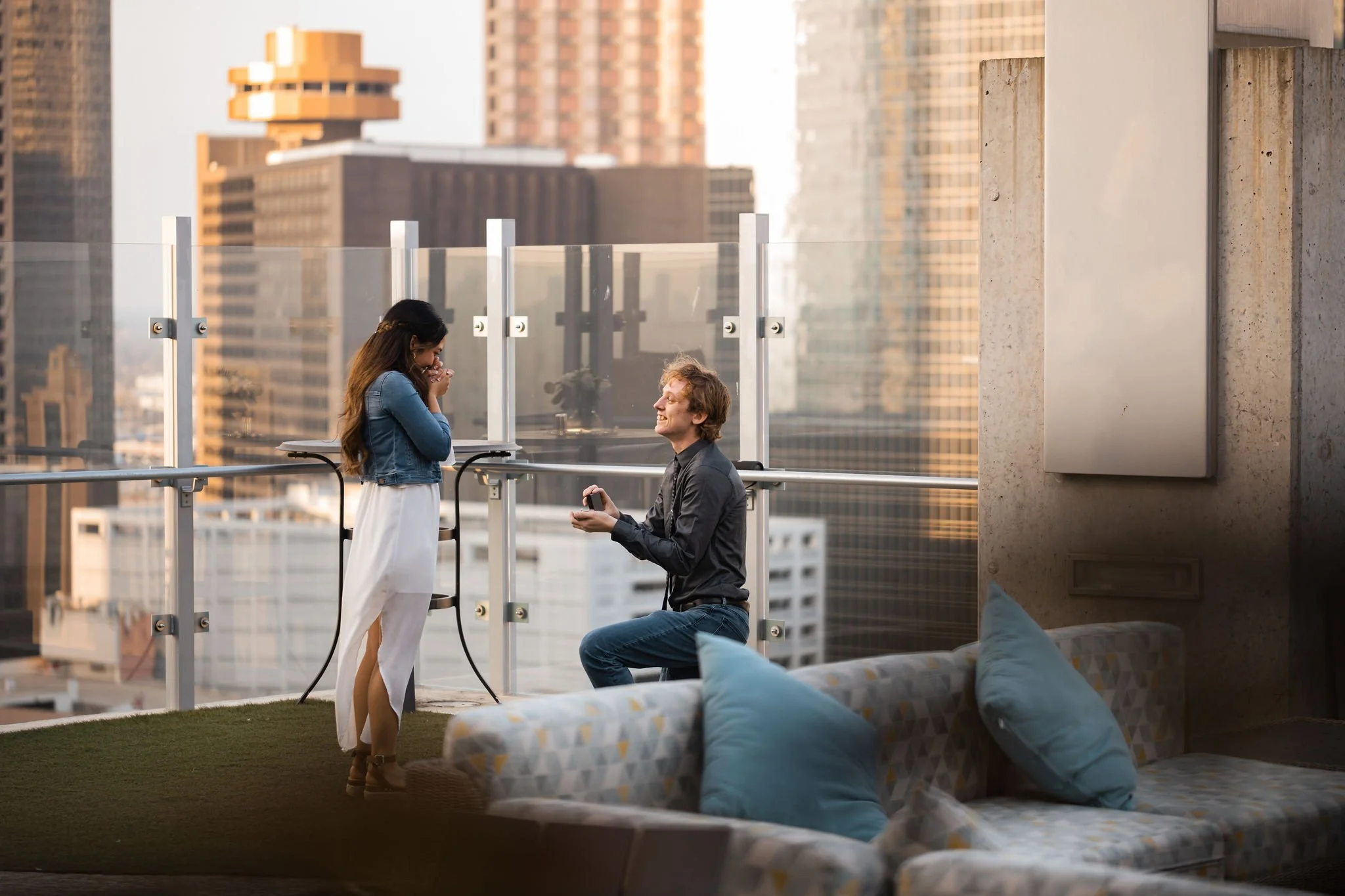 A man is kneeling on one knee proposing to a woman on a balcony with a cityscape background, with a couch and blue pillows visible in the foreground.