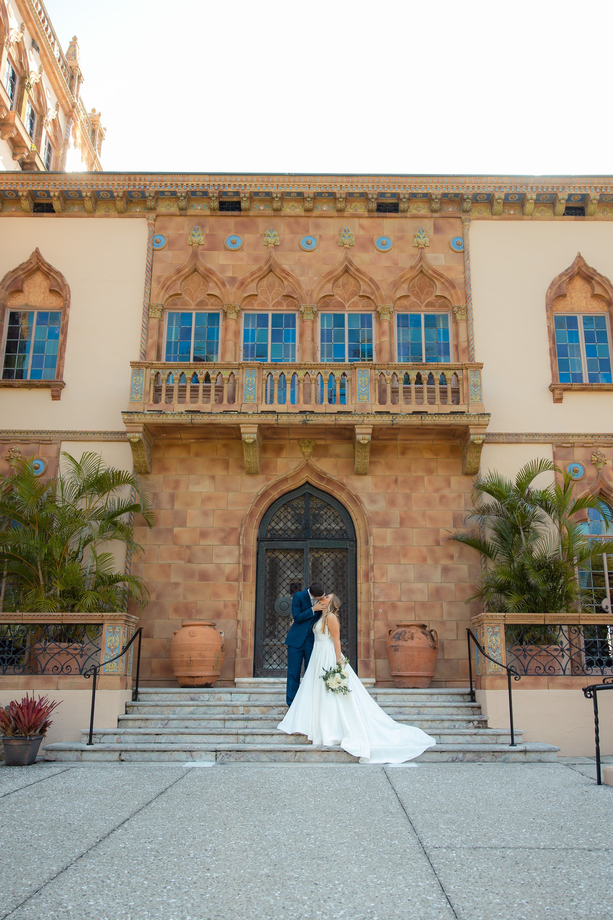 A bride and groom share a kiss on the steps of a historic building with ornate windows and architectural details, surrounded by potted plants.