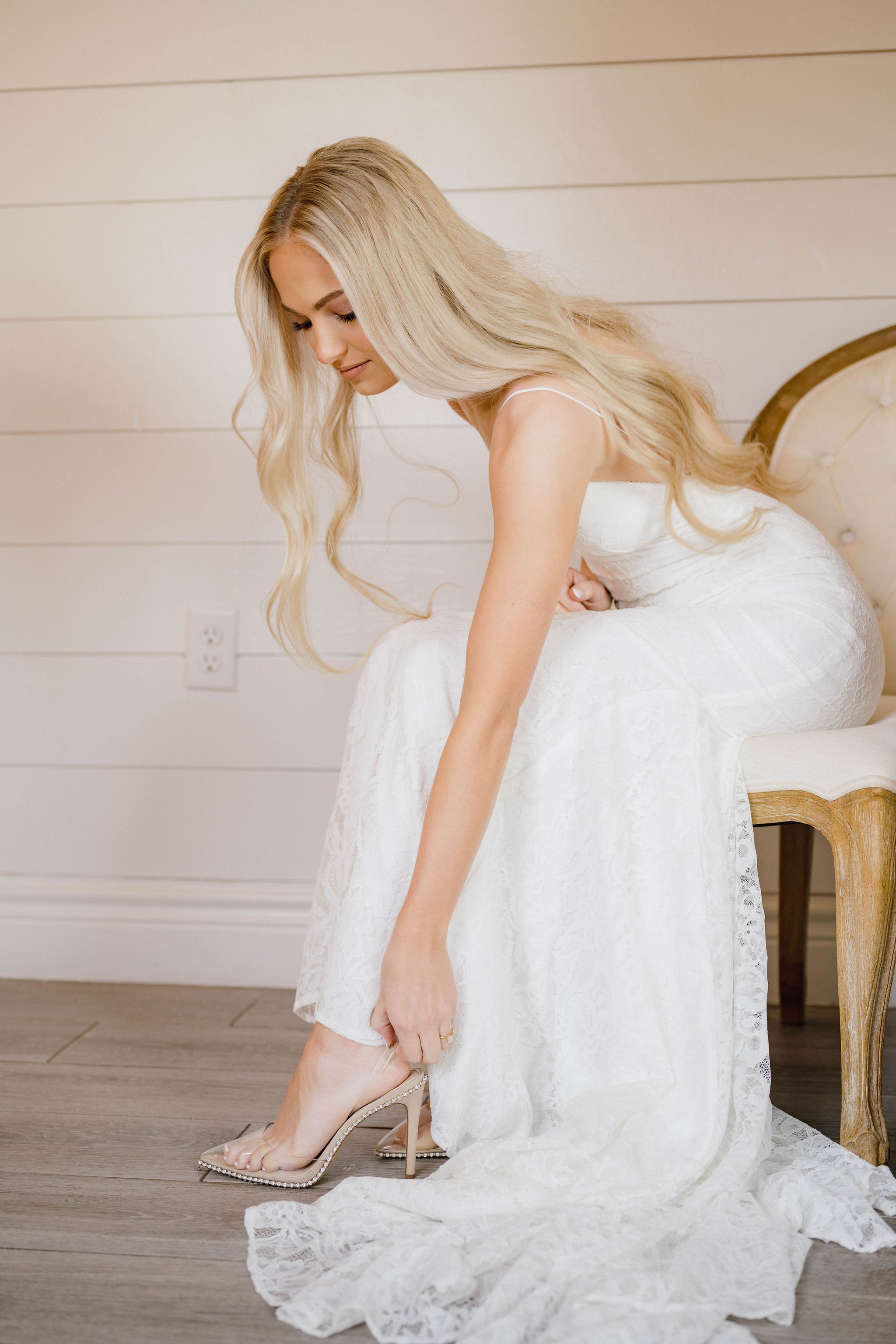 A woman in a white wedding dress sitting on a cream-colored armchair, putting on a clear high-heeled shoe with pearl accents, with a light wood floor and white wall in the background.