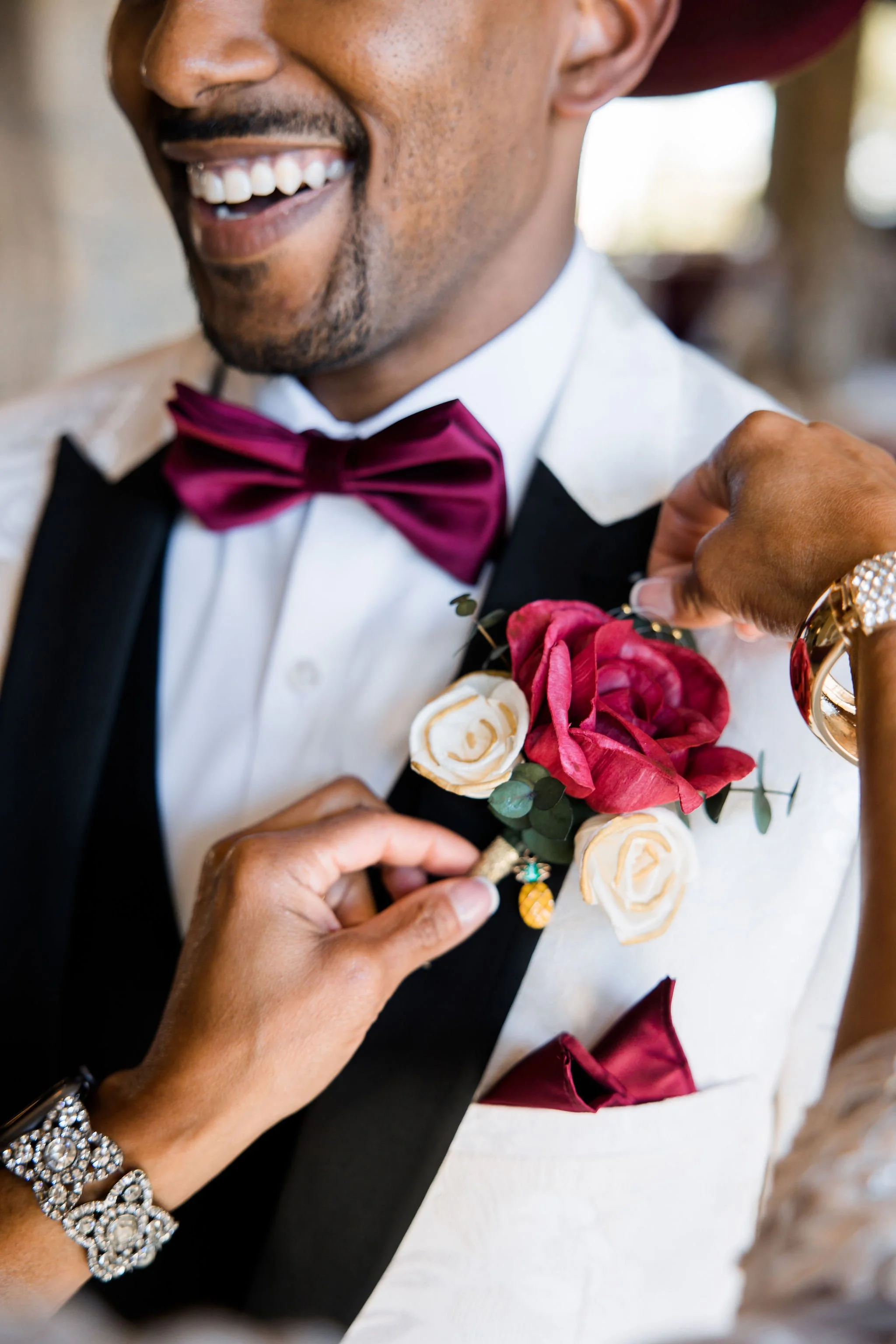 A man in formal attire with a white shirt, black tuxedo, and red bow tie has a boutonniere of red and white roses pinned to his tuxedo; someone is adjusting the boutonniere, and the man is smiling.