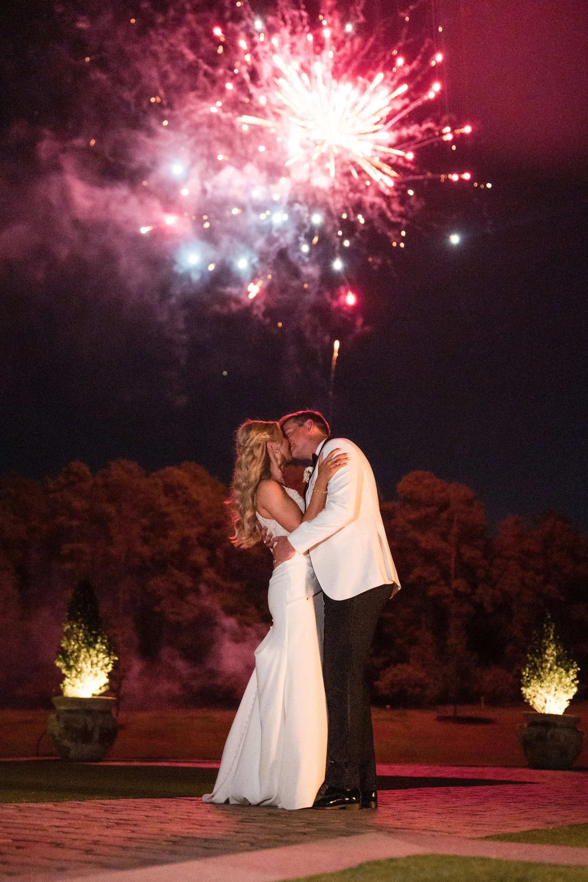 A bride and groom kissing under a fireworks display at night, outside during their wedding celebration.