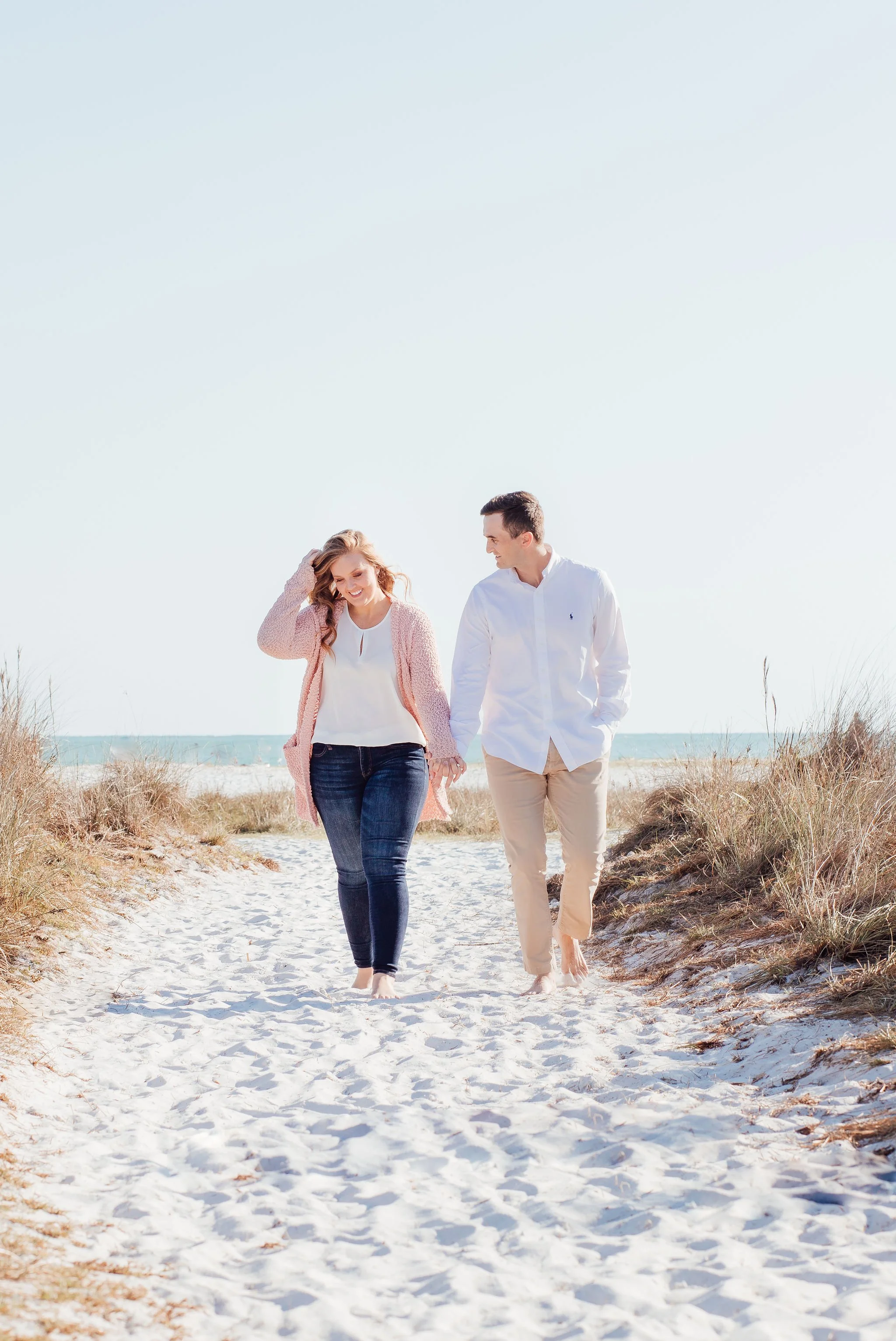 A young couple walking barefoot on a sandy beach pathway, holding hands, with dunes and the ocean in the background on a clear, sunny day.