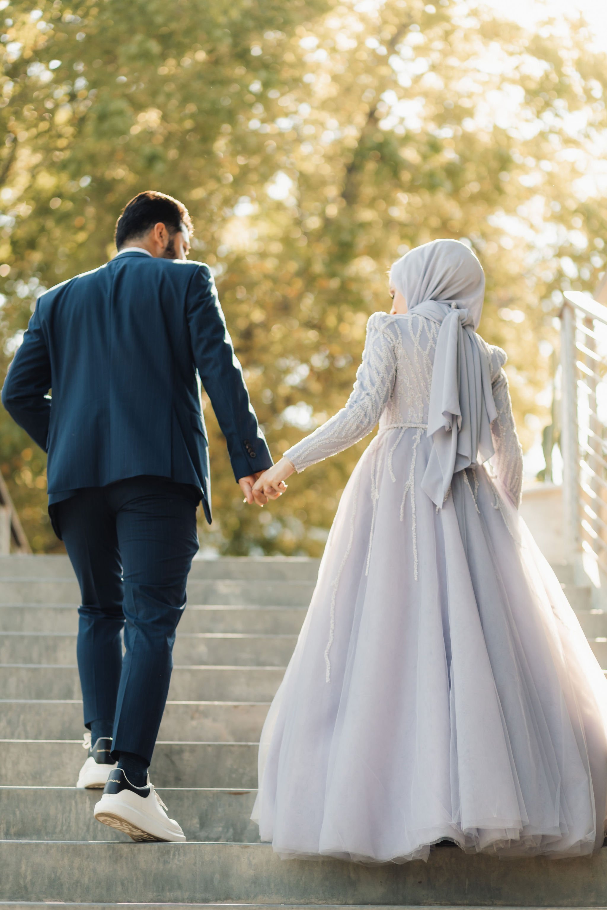 A couple holding hands and walking up outdoor stairs, with a man in a dark suit and a woman in a white gown and hijab, surrounded by trees with sunlight filtering through.