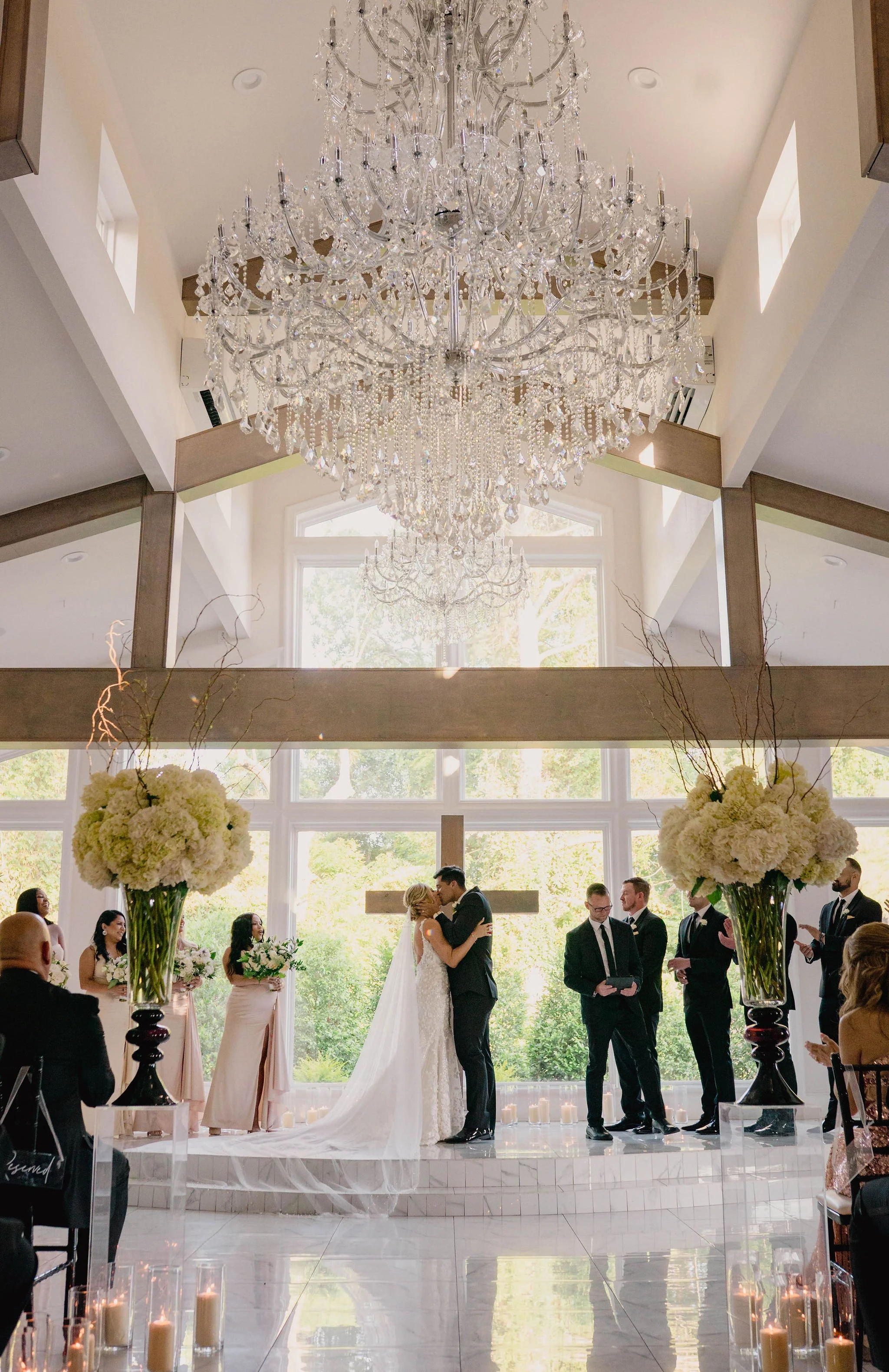 A wedding ceremony taking place in a bright, modern chapel with a large chandelier hanging from the ceiling. The bride and groom are kissing at the altar, surrounded by bridesmaids, groomsmen, and guests, with floral arrangements and candles.