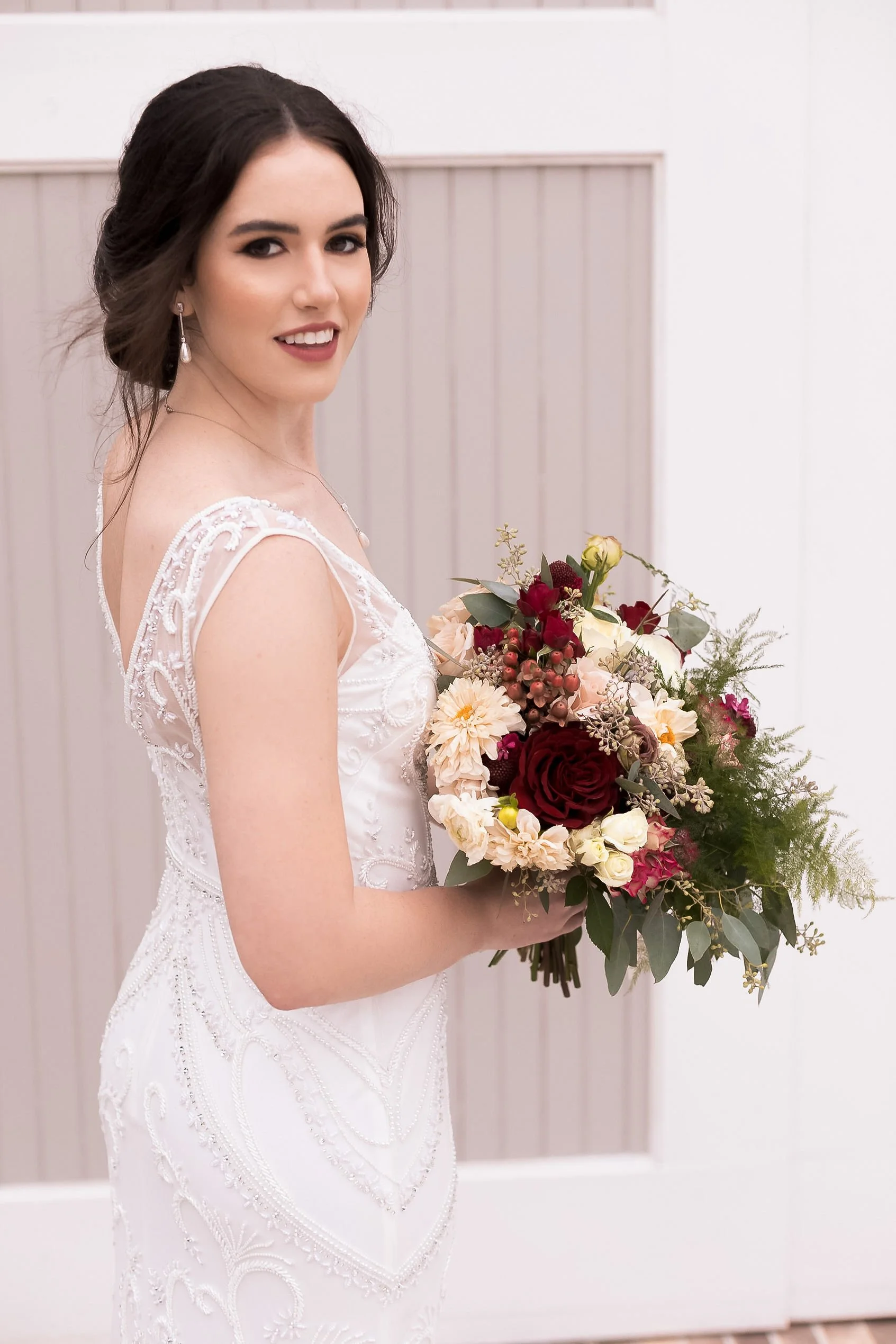 A bride with dark hair in an updo, wearing a white lace wedding dress and pearl earrings, holding a bouquet of red, cream, and pink flowers with greenery, smiling.