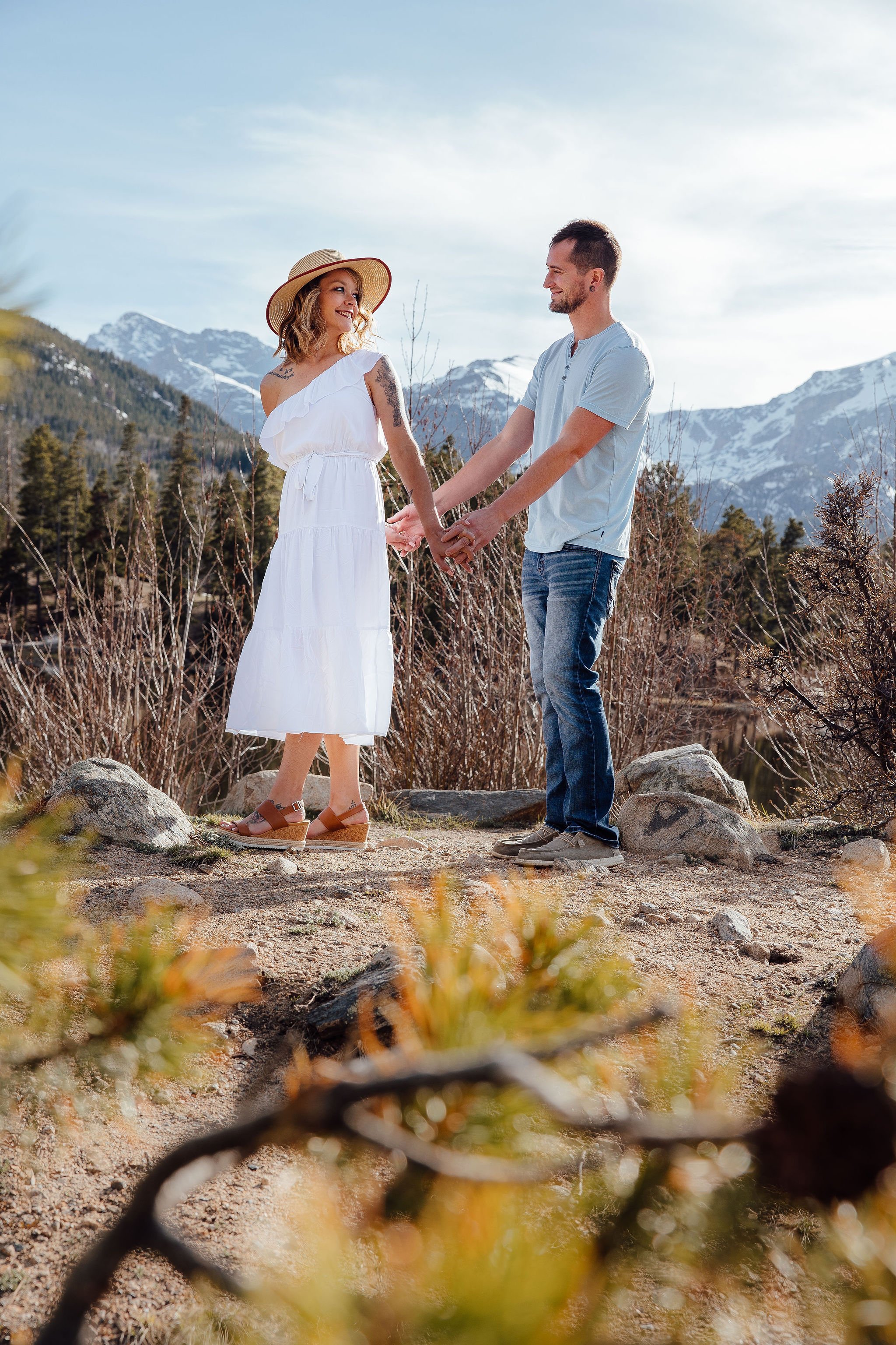 A couple holding hands and smiling at each other outdoors with mountain scenery in the background.