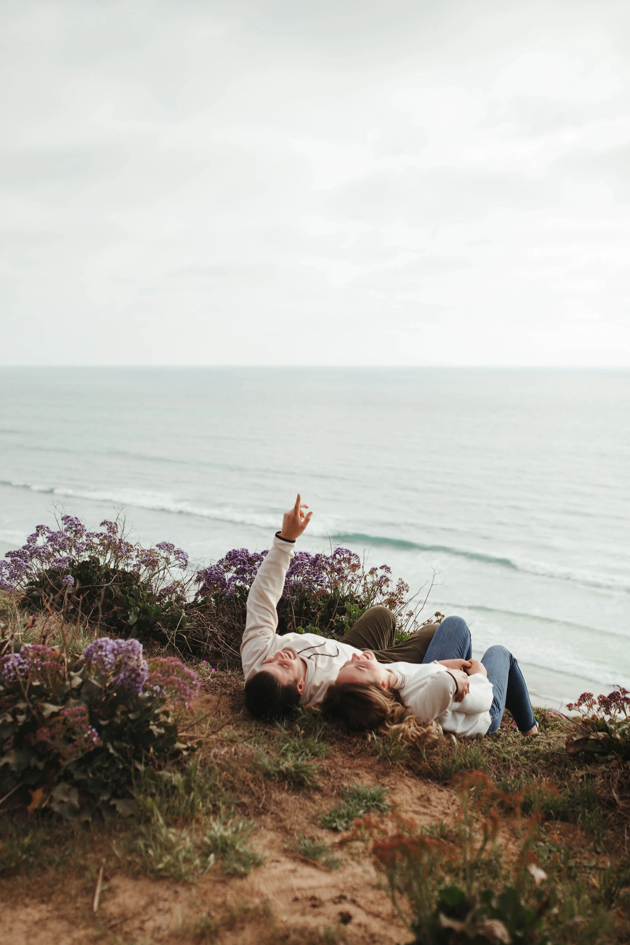 A couple lying on the ground near the shore, surrounded by purple flowers, with the ocean and overcast sky in the background.