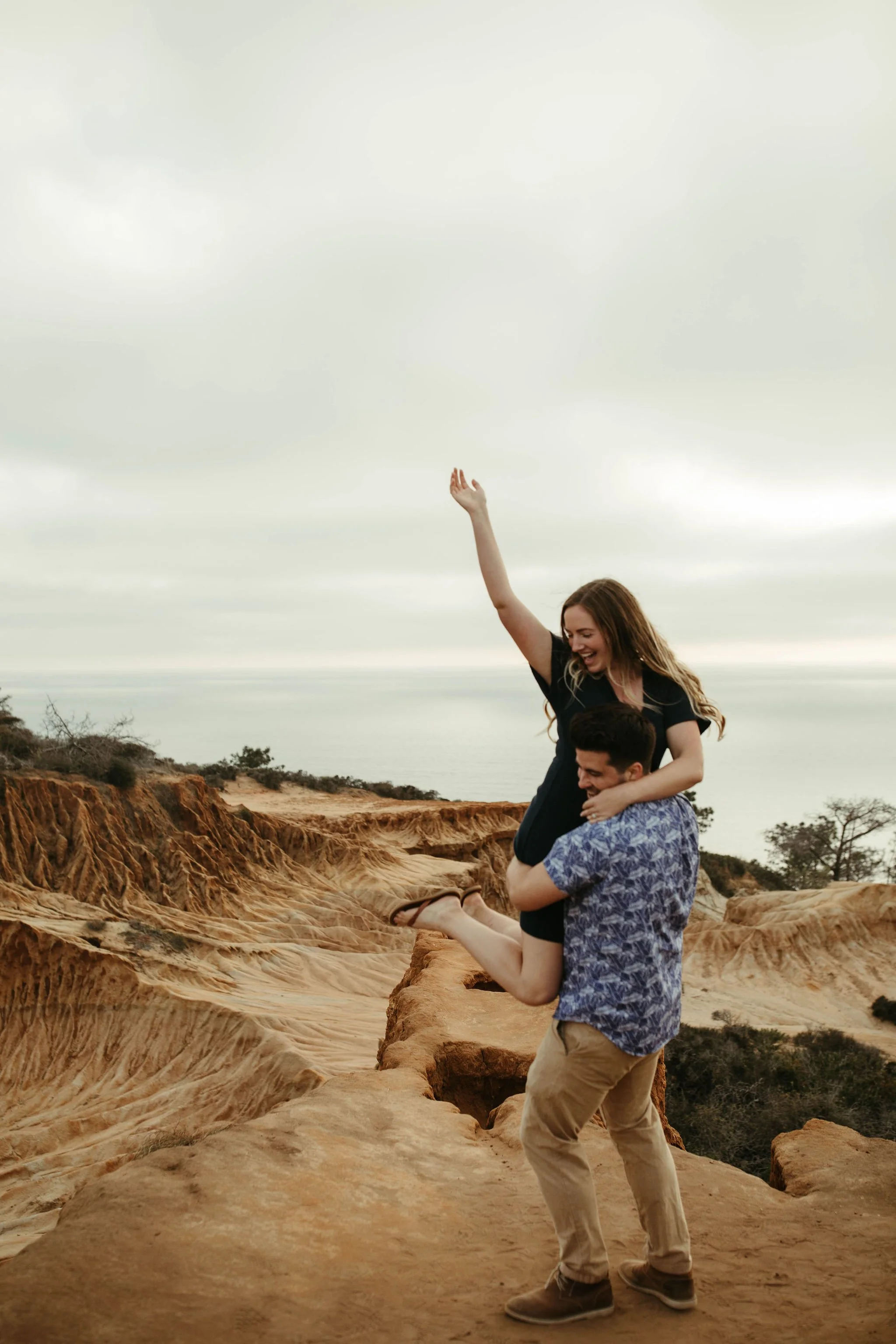 A woman being lifted into the air by a man on a rocky cliff in a desert landscape with cloudy sky.