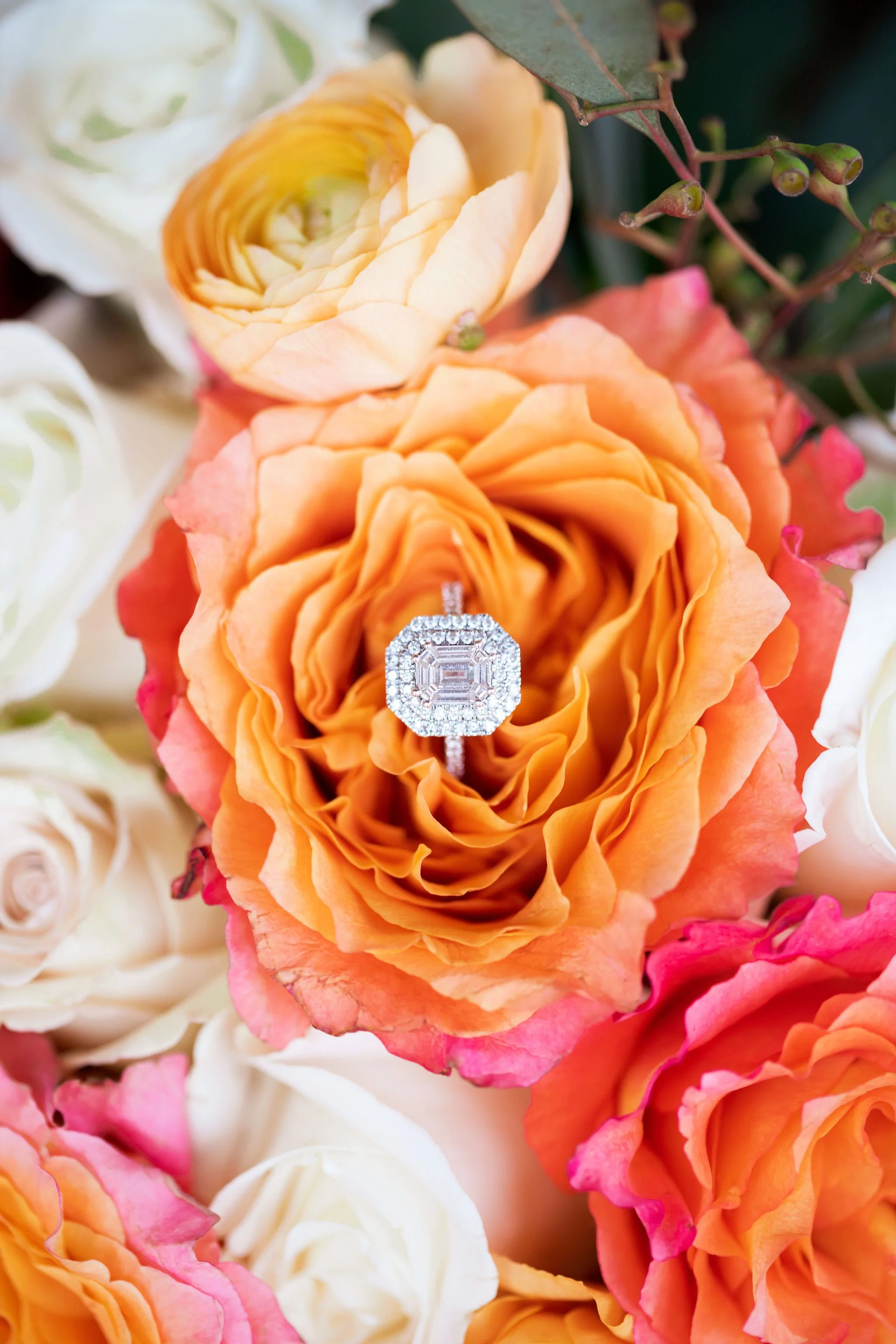 A diamond engagement ring with an emerald-cut center stone surrounded by a halo of smaller diamonds is placed on an orange rose among white and pink roses.