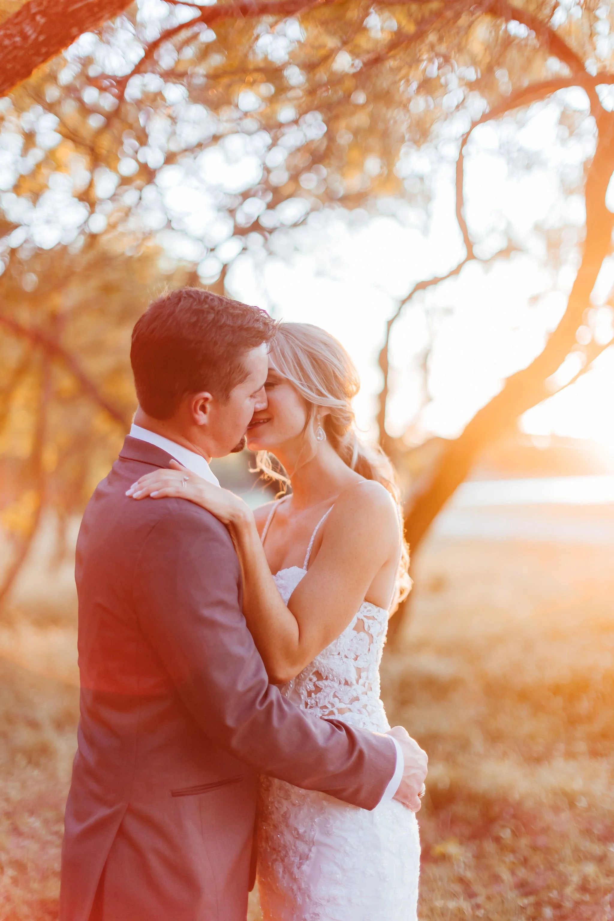 A newlywed couple sharing an intimate moment outdoors during sunset, with autumn trees in the background.